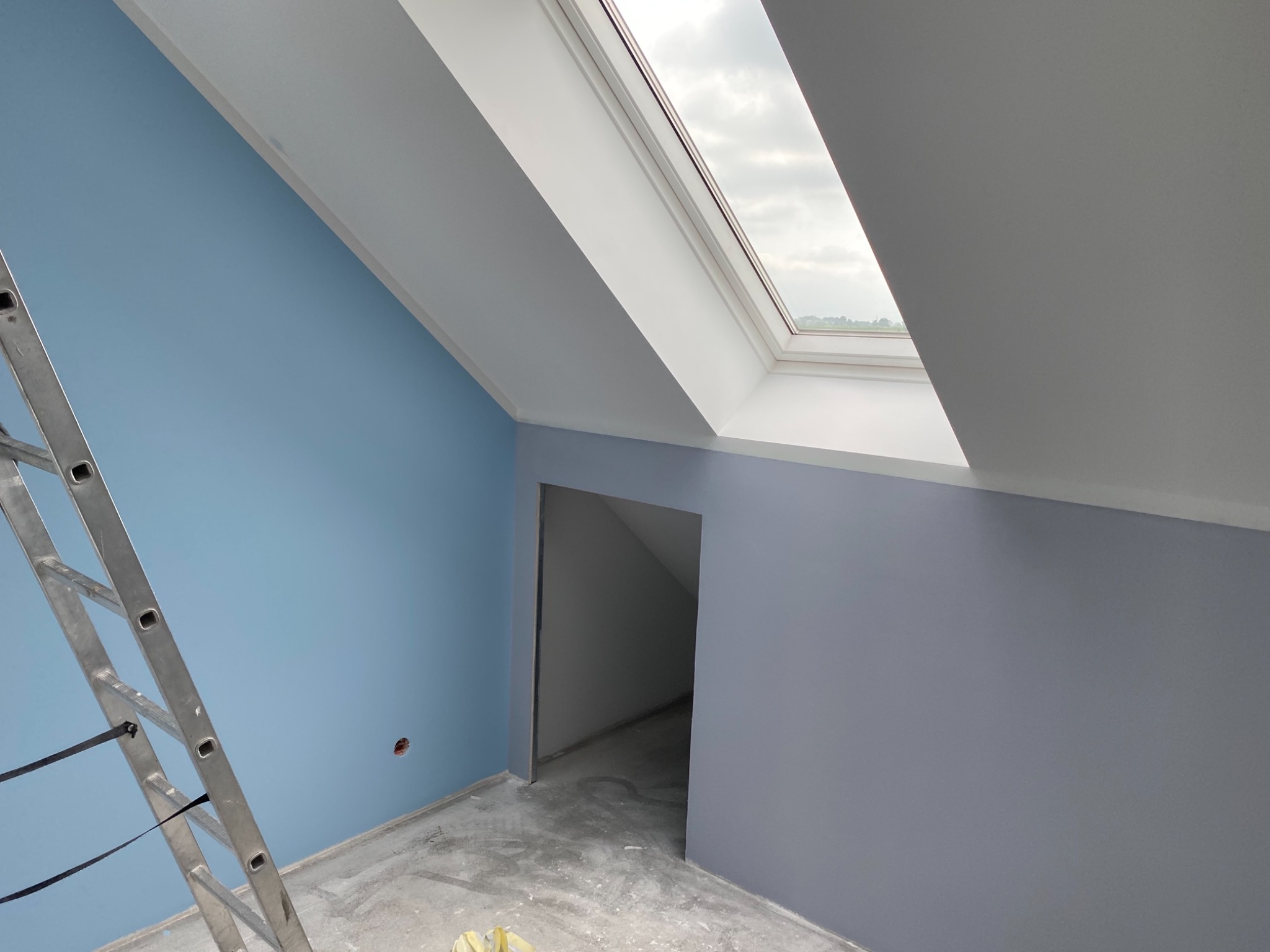 Wnętrze pokoju na poddaszu z pomalowanymi ścianami na niebiesko i szaro, widoczna drabina aluminiowa i okno dachowe.