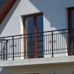 Czarna, metalowa balustrada balkonowa z dekoracyjnymi elementami na tle białej ściany domu z brązowymi oknami i błękitnym niebem.