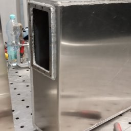 SPAW-TECH Mateusz Pokusa - Spawana, metalowa obudowa z otworem, widoczne spawy. Na stole spawalniczym, obok butelka wody. Detal wykonany z blachy nierdzewnej.