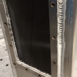 SPAW-TECH Mateusz Pokusa - Szczegółowe zbliżenie na precyzyjne spawy na metalowej konstrukcji. Widoczne regularne ściegi spawalnicze i otwory montażowe. W tle narzędzia i butelka wody.