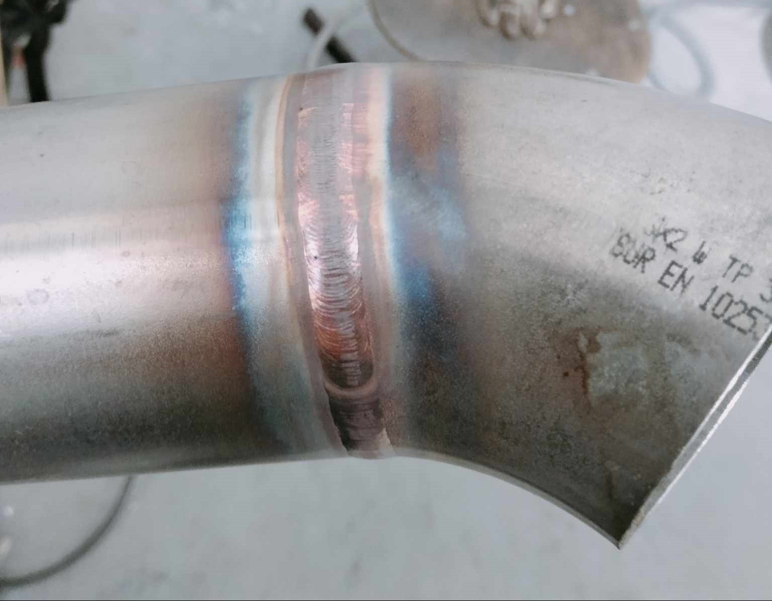 Spoina na zagiętym elemencie rury ze stali nierdzewnej, widoczne ślady po procesie łączenia i oznaczenia normy EN 10257.