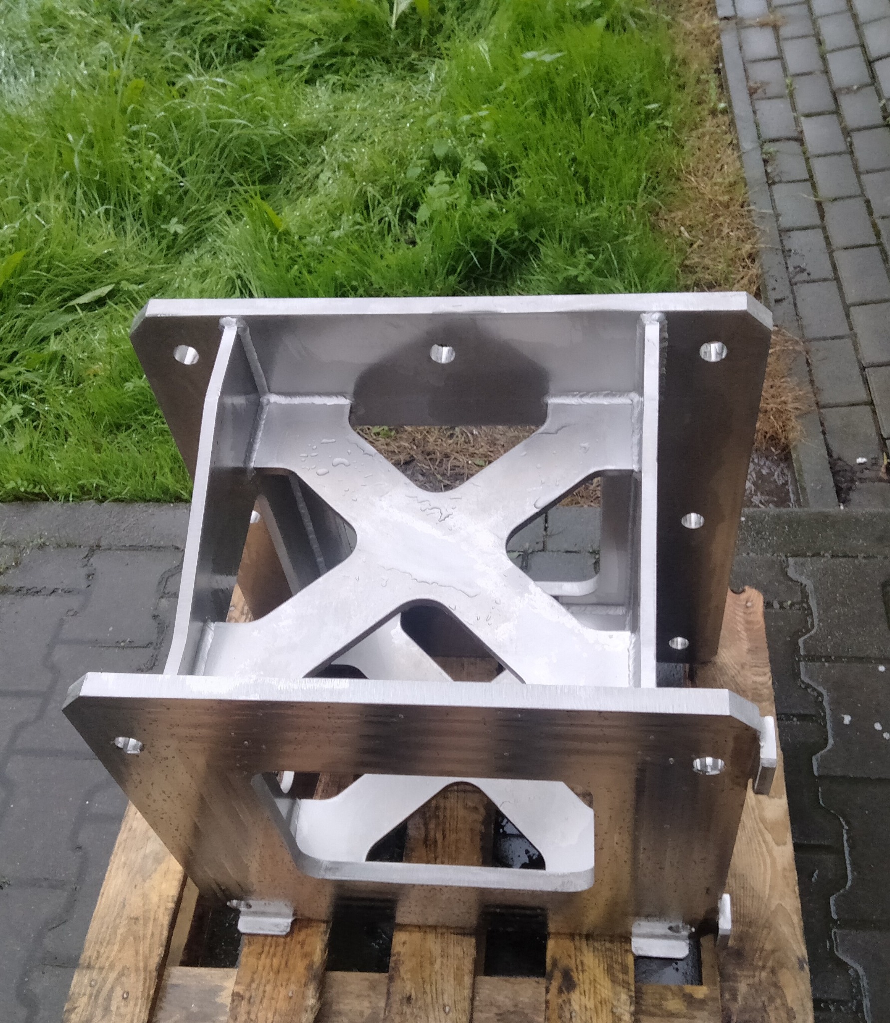 Metalowa konstrukcja spawana z otworami, na drewnianej palecie, z widocznymi śladami obróbki i spawania. Tło stanowi trawa i kostka brukowa.