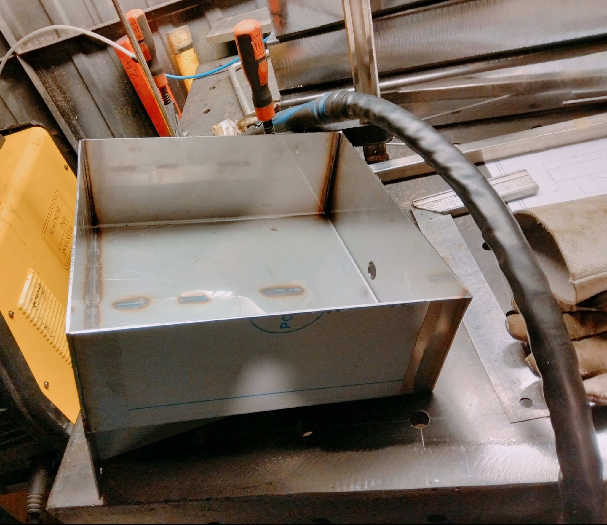 Metalowa skrzynka z widocznymi spawami, leżąca na stole warsztatowym obok spawarki i narzędzi. Detal z bliska, ukazujący precyzję wykonania i estetykę spoin.