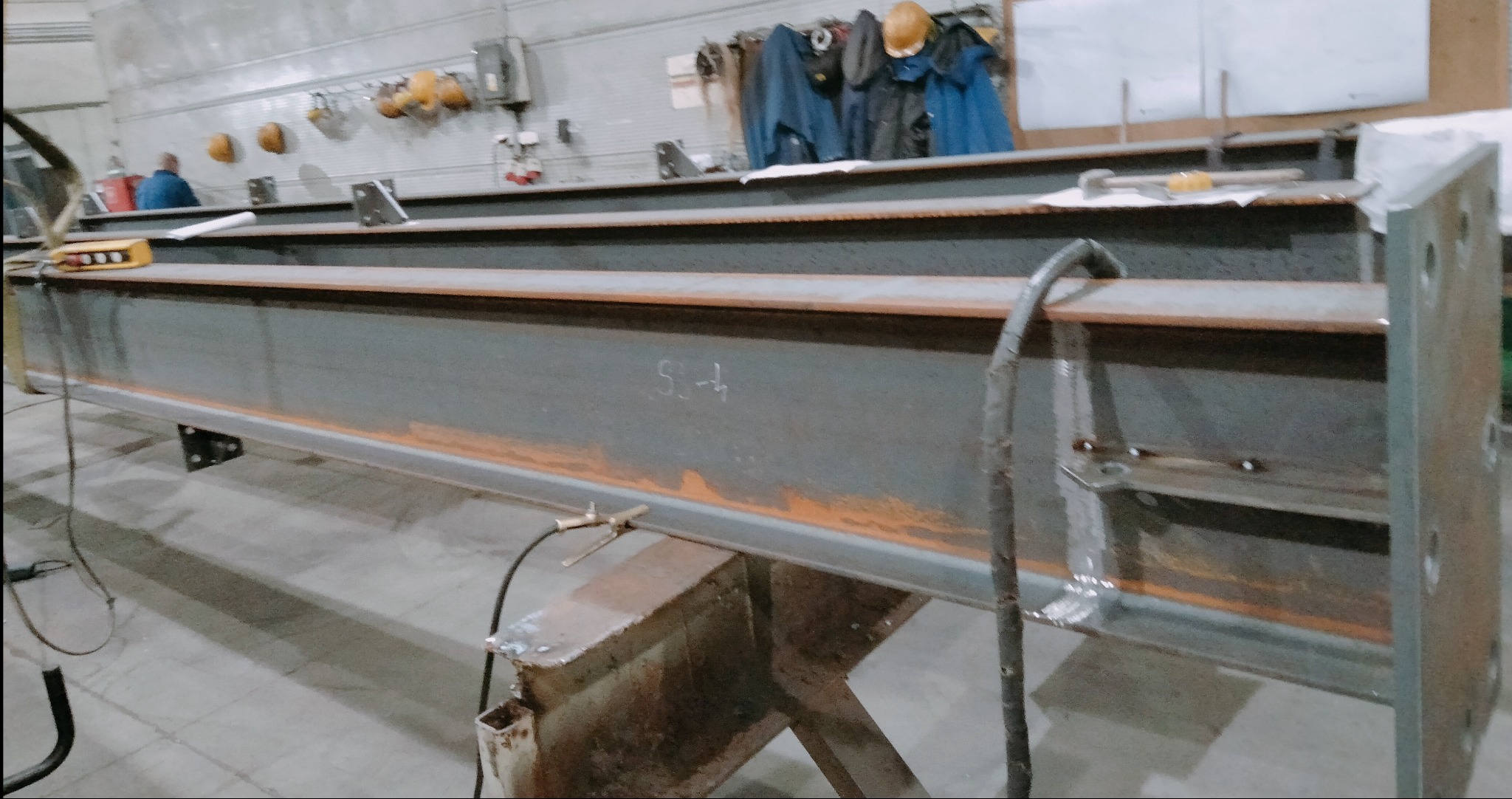 Długa, surowa belka stalowa na warsztacie, z widocznymi śladami spawania i oznaczeniami. W tle narzędzia i ubrania robocze, co sugeruje etap obróbki metalu.