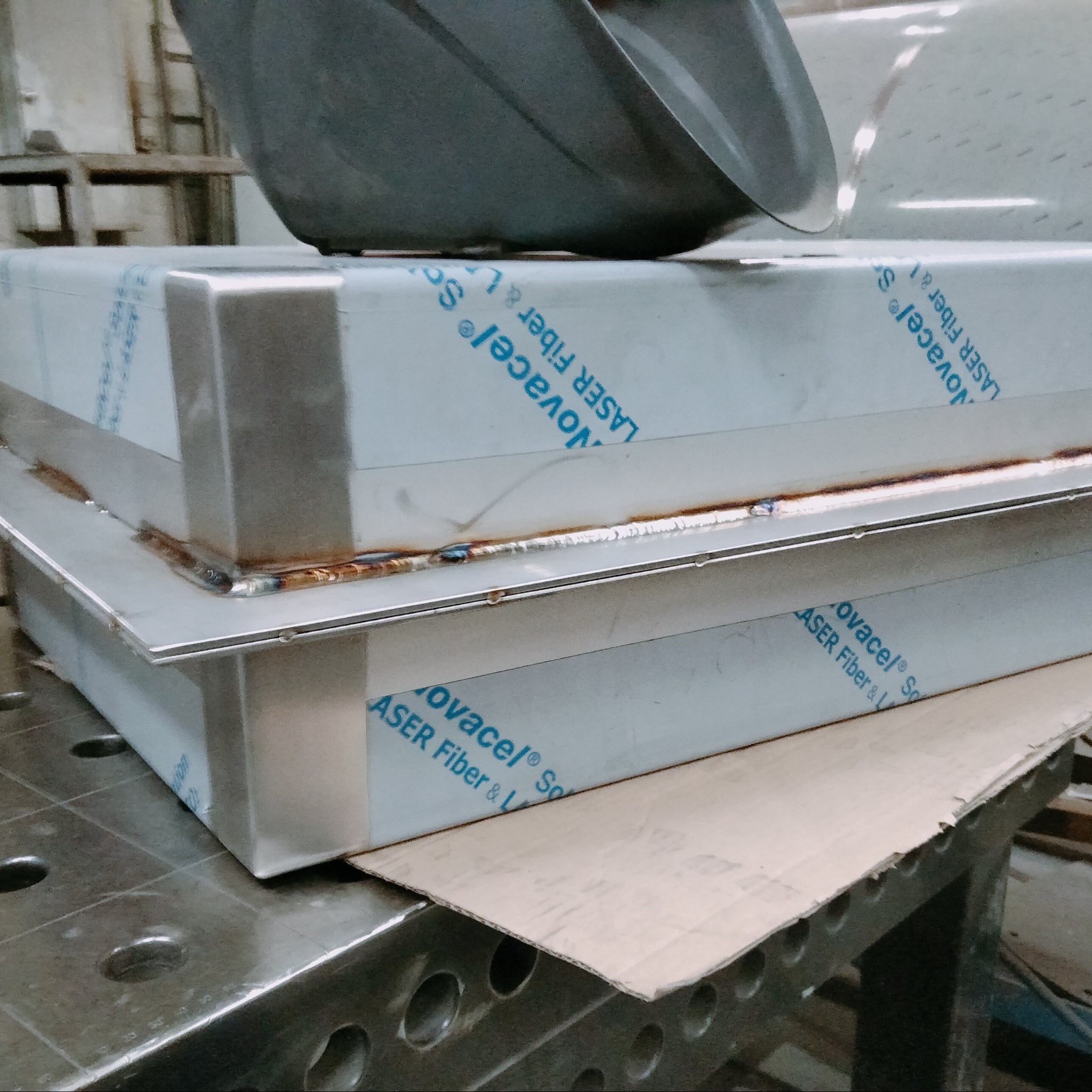 Spawanie narożników metalowych elementów z folią ochronną, widoczne spoiny na krawędziach. Elementy leżą na perforowanym stole spawalniczym.