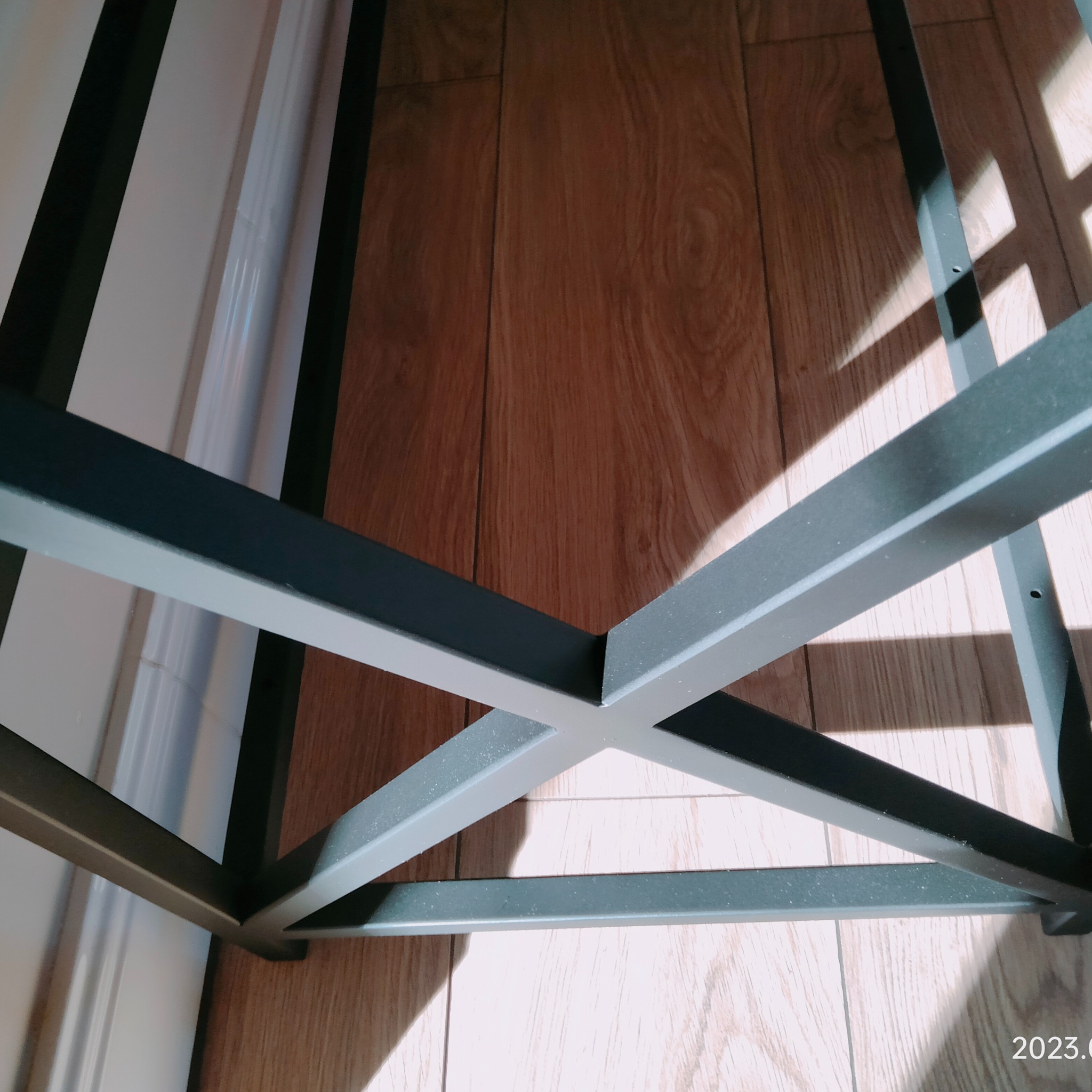 Metalowa konstrukcja podstawy stołu z profili kwadratowych, spawana, pomalowana na szaro, widoczna na tle drewnianej podłogi i białej ściany. Ujęcie z dołu, z bliska.