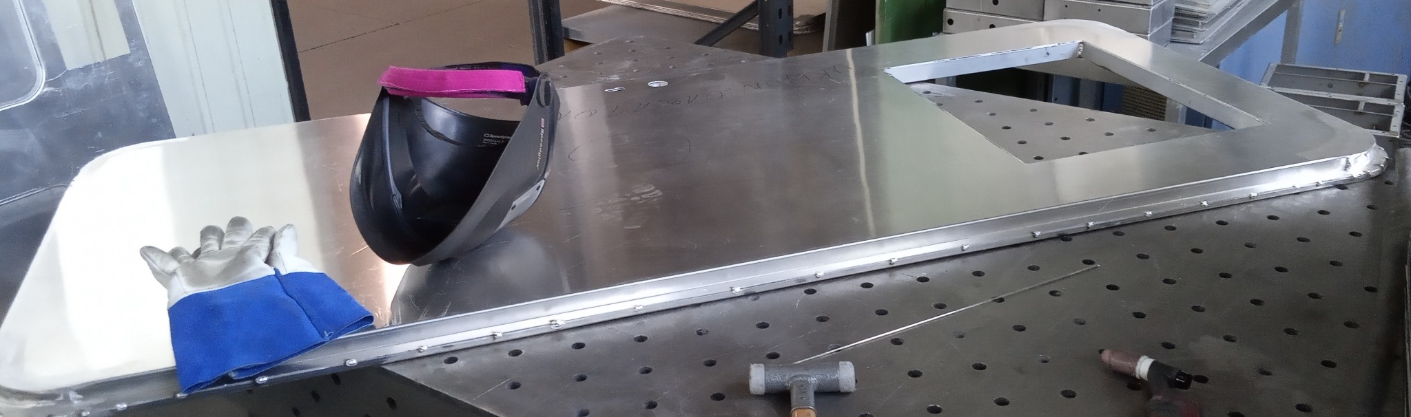 Metalowa konstrukcja spawana na stole spawalniczym, obok maska spawalnicza z różową opaską, rękawice i młotek. Widoczne ślady spawania.