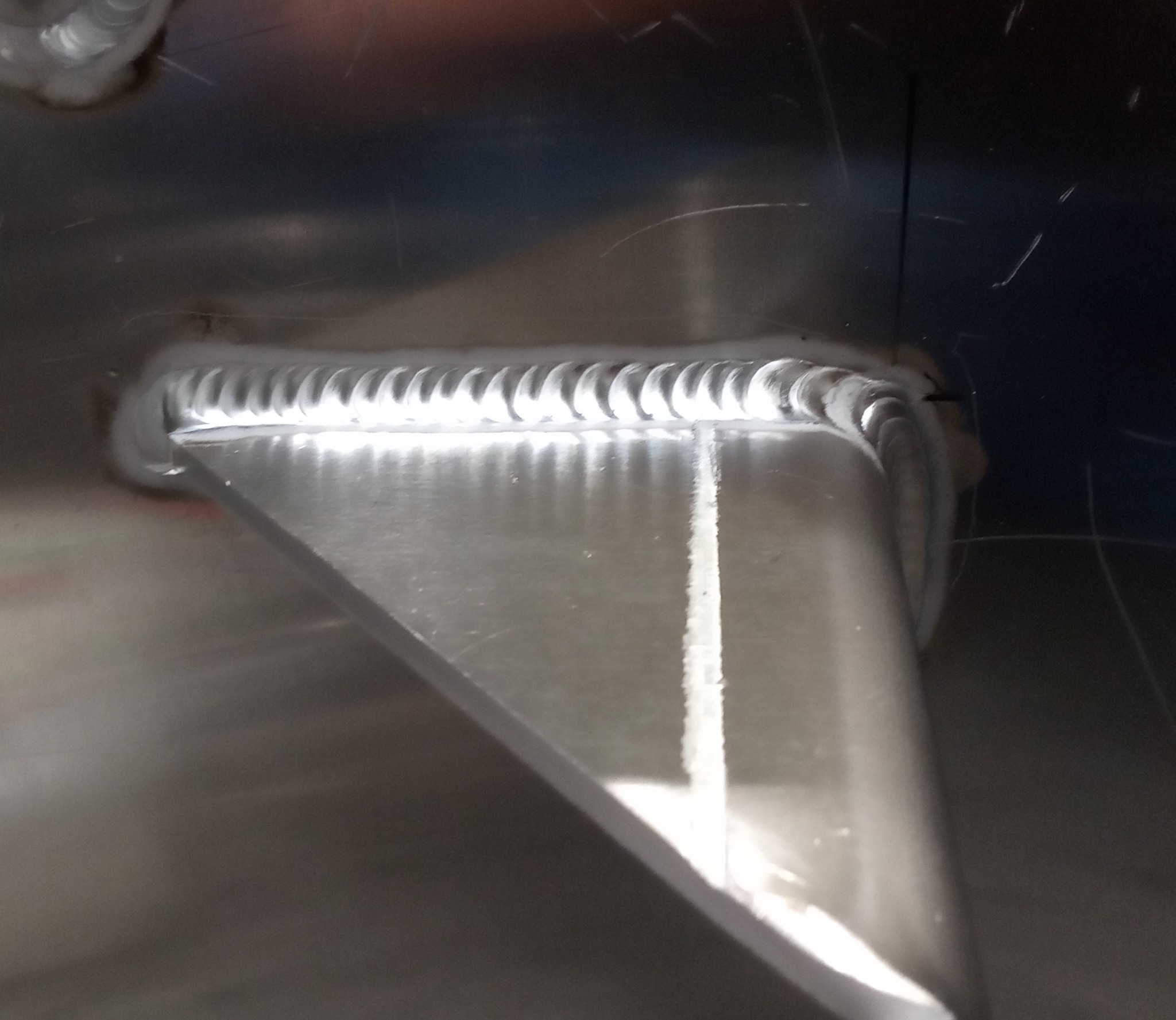 Precyzyjne spawy łączące metalowe elementy pod kątem, widoczne regularne ślady spawania na gładkiej powierzchni. Detal z bliska, srebrzyste odcienie metalu.