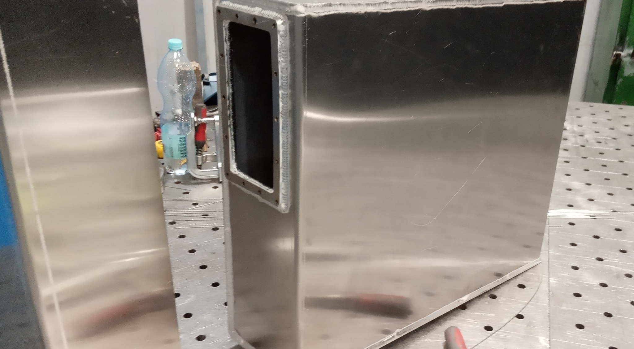 Spawana, metalowa obudowa z otworem, widoczne spawy. Na stole spawalniczym, obok butelka wody. Detal wykonany z blachy nierdzewnej.