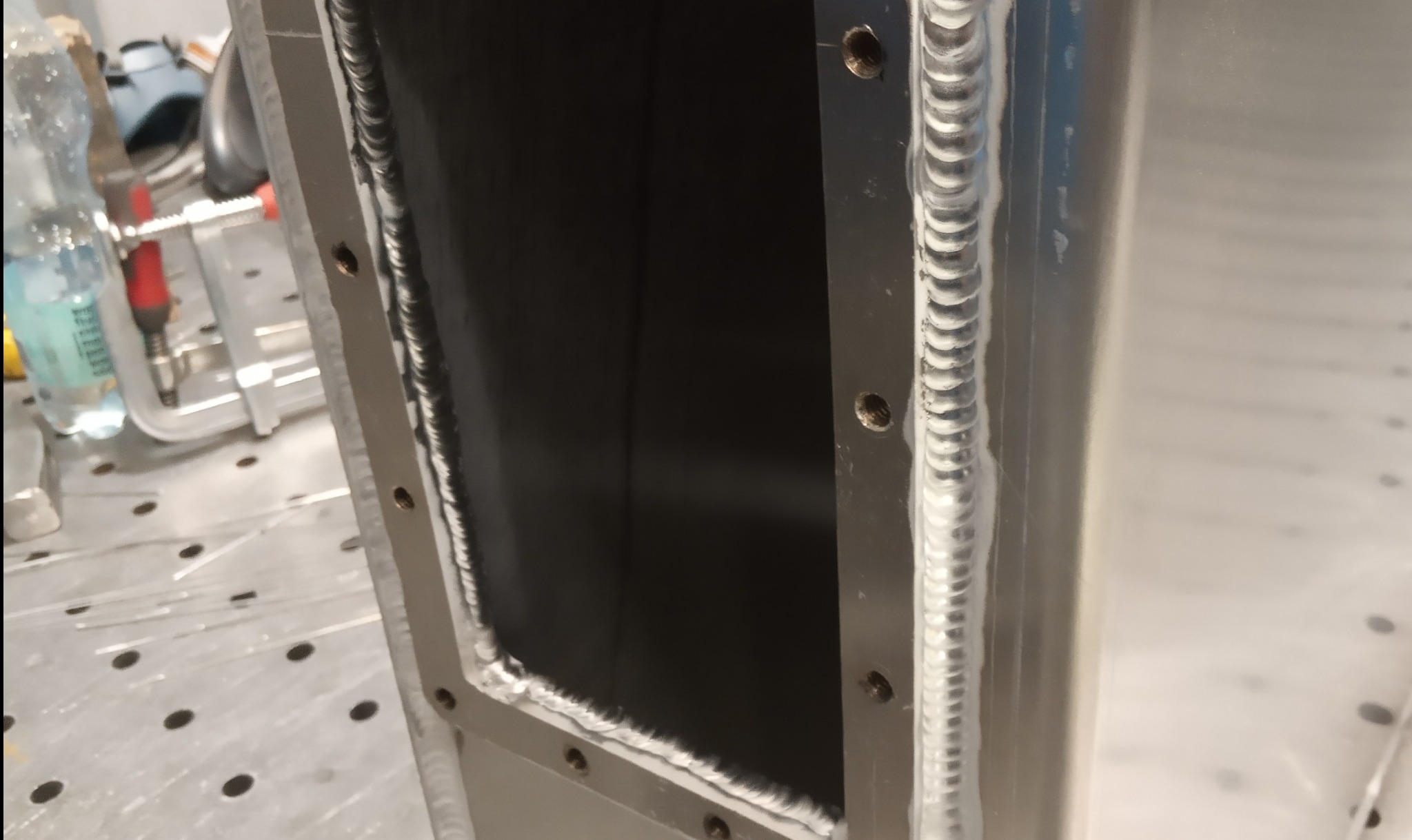 Szczegółowe zbliżenie na precyzyjne spawy na metalowej konstrukcji. Widoczne regularne ściegi spawalnicze i otwory montażowe. W tle narzędzia i butelka wody.