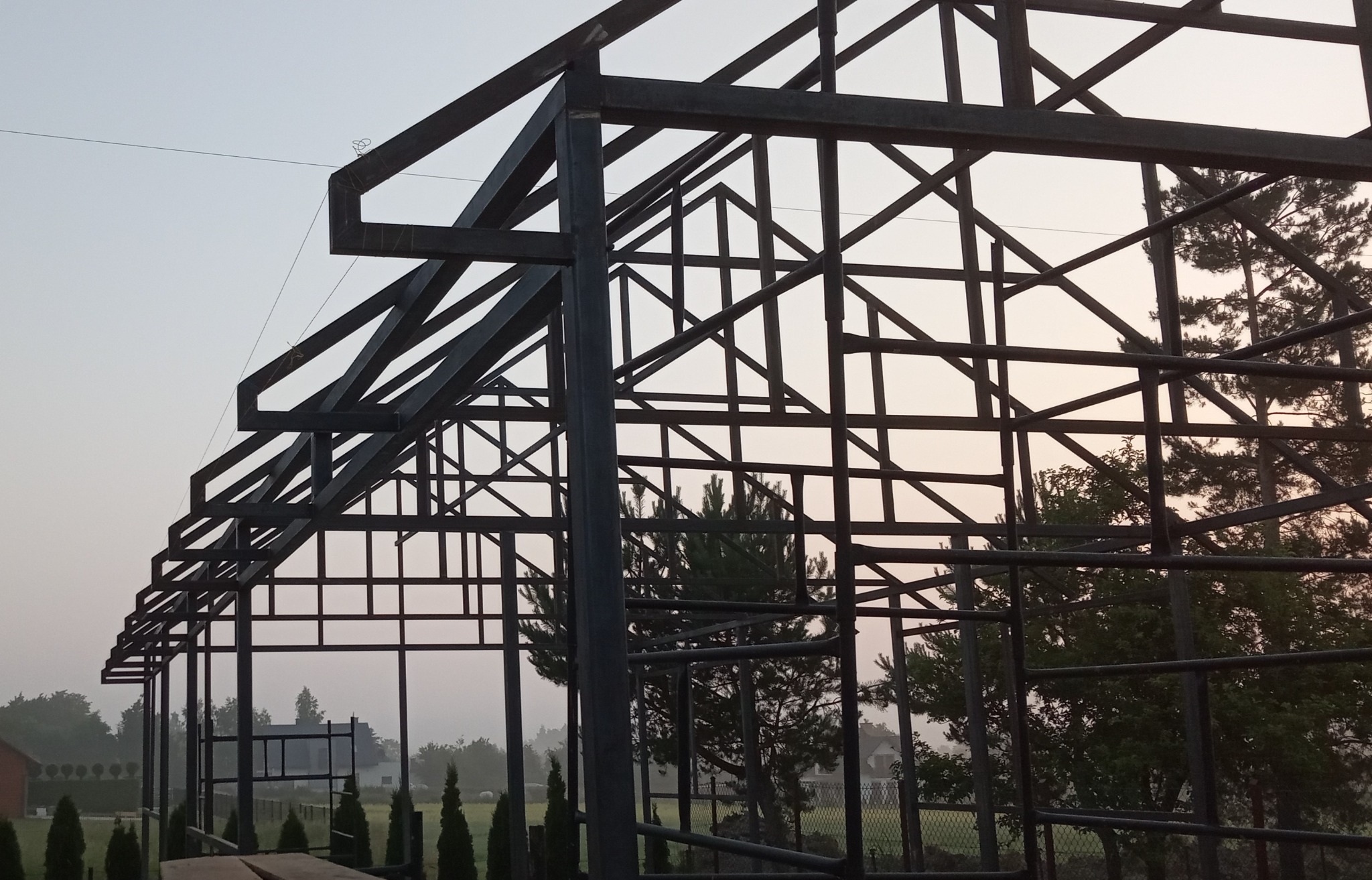 Konstrukcja metalowa w trakcie budowy, widok z dołu na tle drzew i nieba o zmierzchu. Elementy spawane, surowy metal, widoczne łączenia.