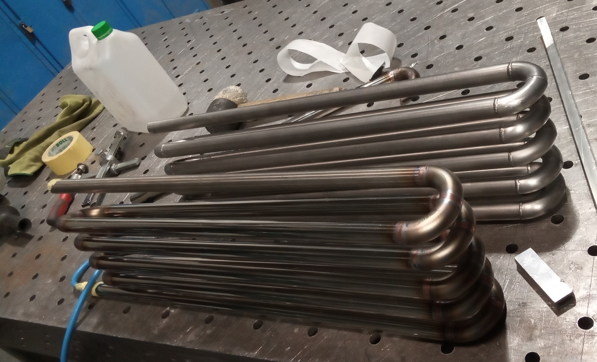 Zespawane rury ze stali nierdzewnej ułożone na perforowanym stole warsztatowym, widoczne ślady spawania, obok narzędzia i akcesoria.
