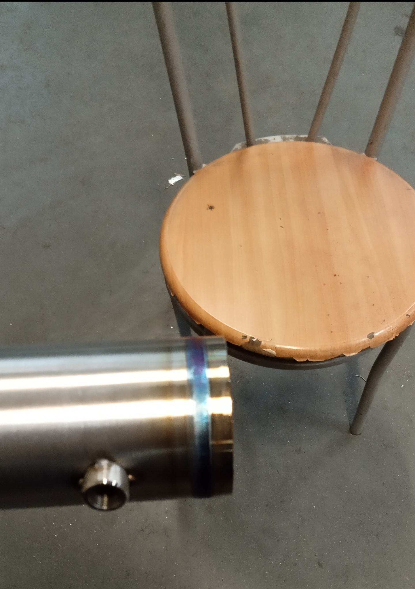 Błyszcząca, metalowa rura z małym otworem, leżąca obok krzesła z drewnianym siedziskiem na szarym tle. Widoczne ślady spawania.