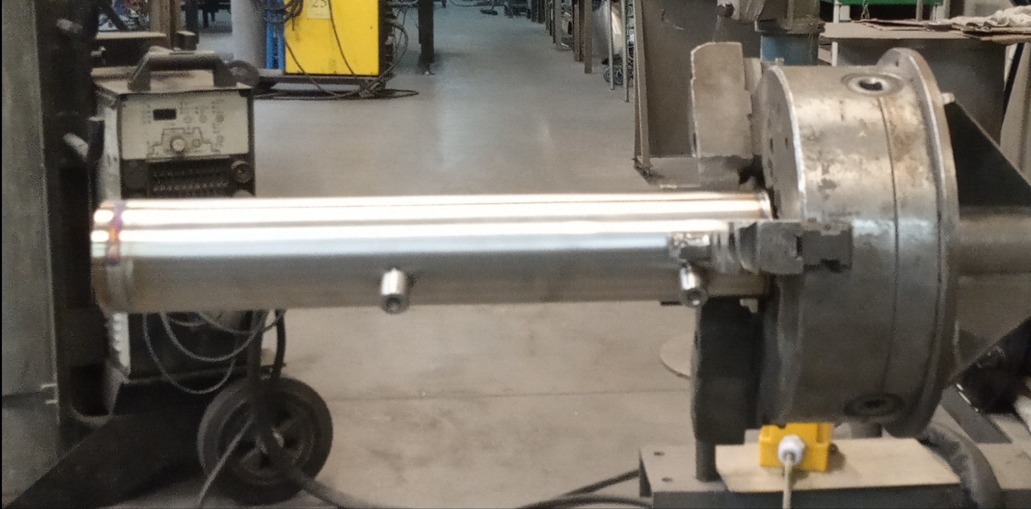 Polerowana rura w uchwycie tokarskim, widoczne ślady spawania. W tle spawarka na kółkach i inne urządzenia warsztatowe. Precyzyjna obróbka metalu.