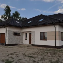 Nowoczesny dom jednorodzinny z grafitową dachówką, elewacją w odcieniach bieli i beżu oraz fragmentami wykończonymi kamieniem dekoracyjnym, widoczny z zewnątrz, na działce przed domem widoczny piasek.