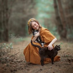 Paulina Gruca - Uśmiechnięta blondynka w musztardowej sukience z wiankiem na głowie, siedząca na leśnym poszyciu i tuląca czarno-białego kota, obok stoi czarny kot z obrożą.