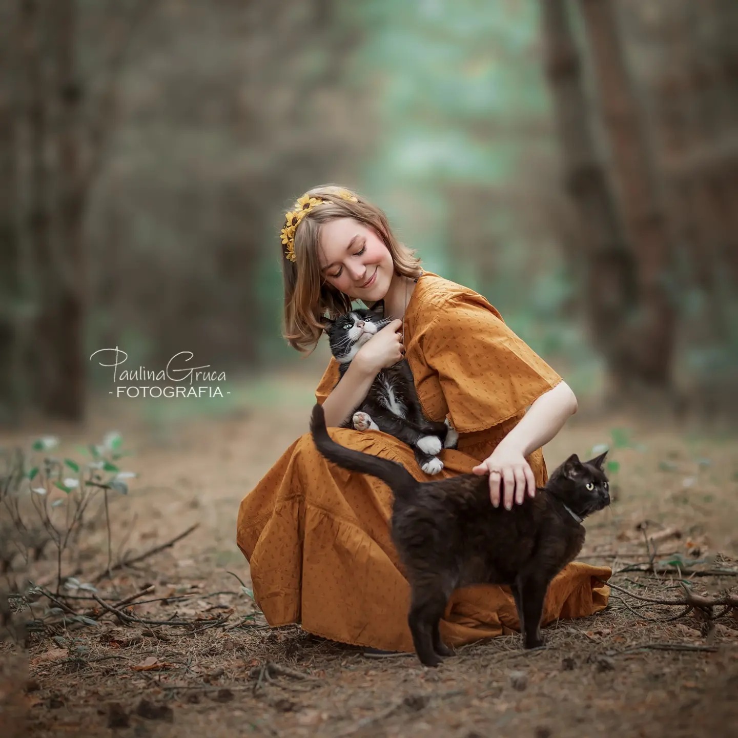 Uśmiechnięta blondynka w musztardowej sukience z wiankiem na głowie, siedząca na leśnym poszyciu i tuląca czarno-białego kota, obok stoi czarny kot z obrożą.