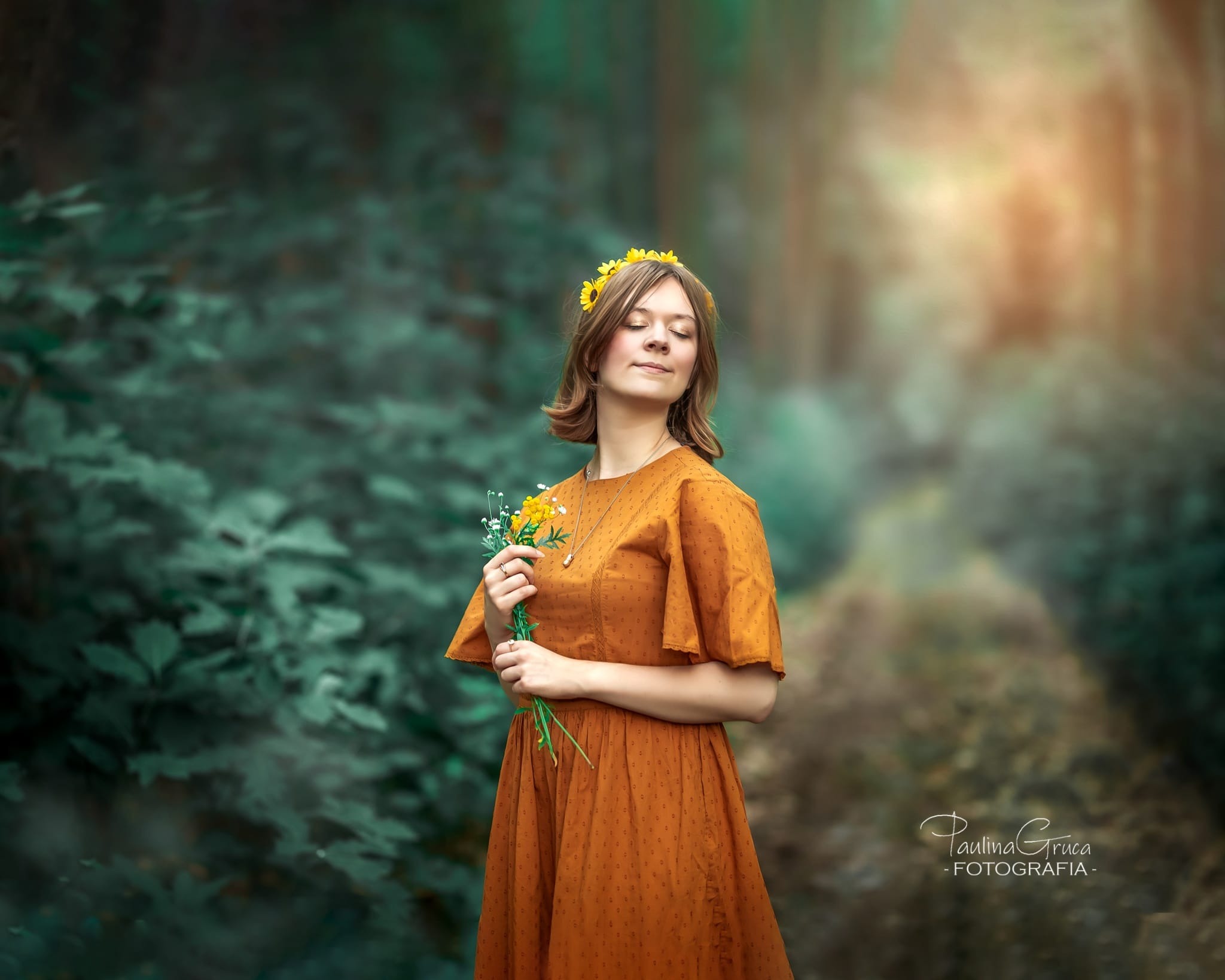Portret młodej kobiety z zamkniętymi oczami, w pomarańczowej sukience i wianku z żółtych kwiatów, trzymającej bukiecik polnych kwiatów na tle leśnej scenerii.