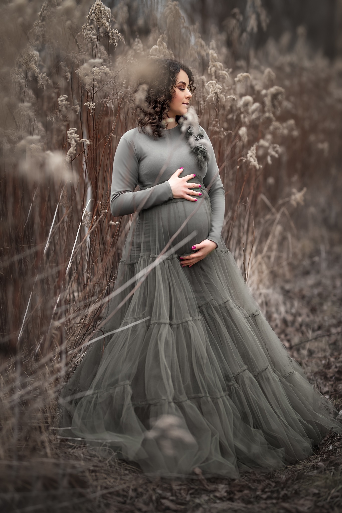Kobieta w ciąży w długiej, szarej sukni z tiulu, pozująca w plenerze na tle uschniętych traw i roślin, z rękami ułożonymi na brzuchu, patrząca w bok.