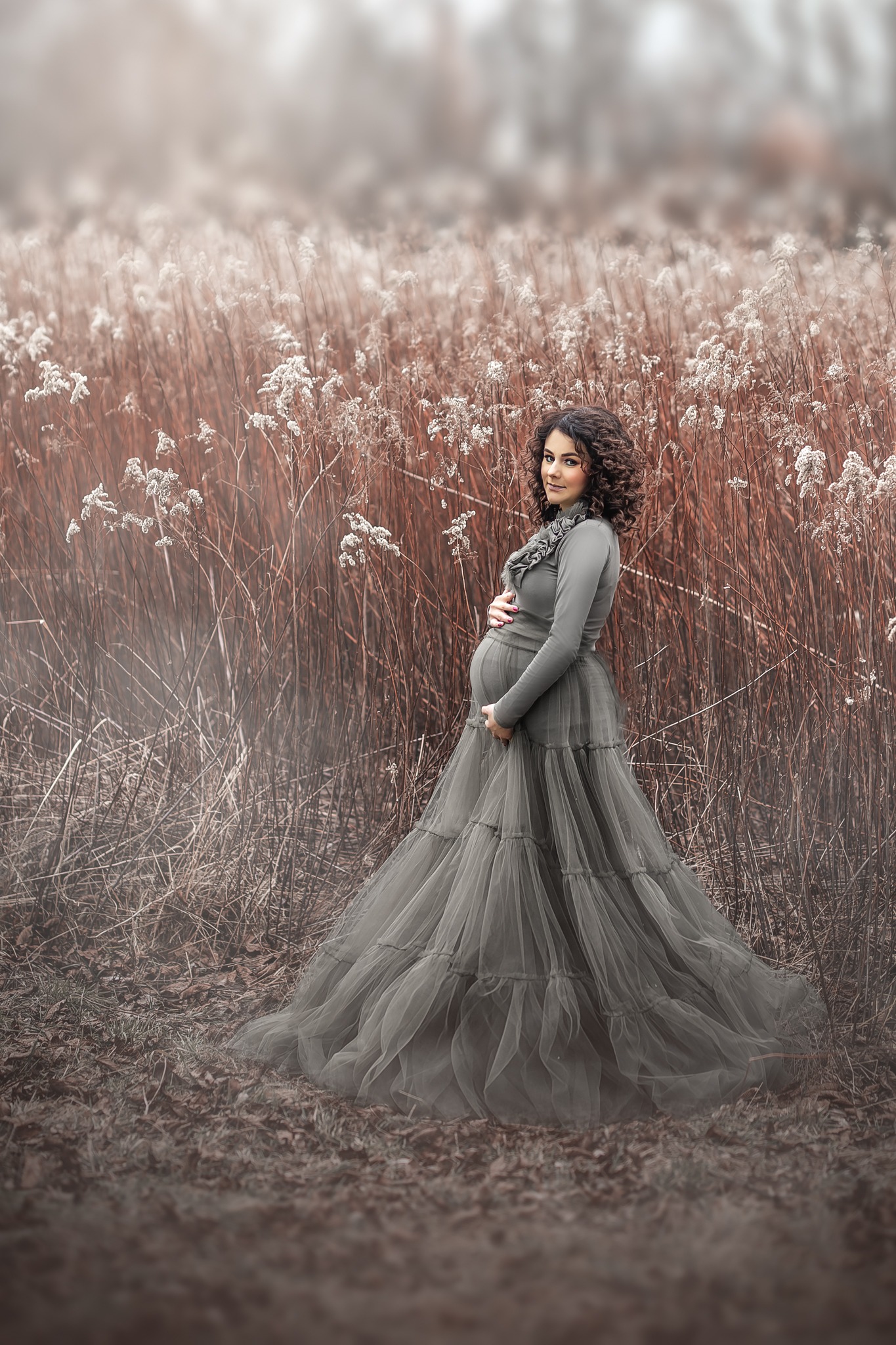 Kobieta w ciąży, ubrana w długą, szarą suknię z tiulu, pozuje na tle wysokich, suchych traw i delikatnych, białych kwiatów, trzymając ręce na brzuchu. Uwagę przyciągają jej kręcone włosy...