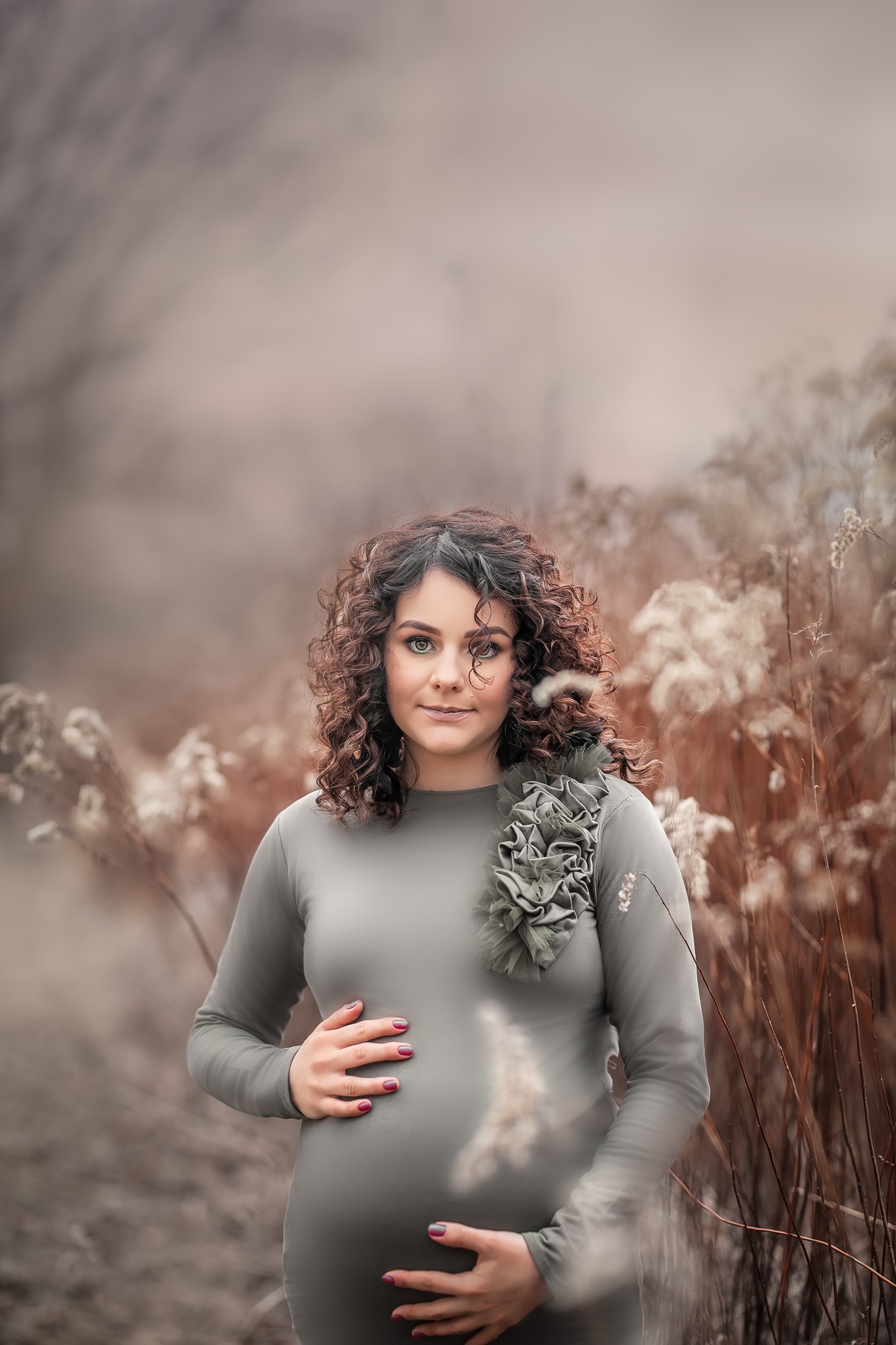 Kobieta w zaawansowanej ciąży, ubrana w dopasowaną szarą sukienkę z ozdobnym kwiatem, pozuje na tle wyschniętej roślinności w plenerze, trzymając dłonie na brzuchu.