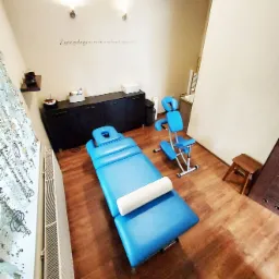 Jasne wnętrze gabinetu z niebieskim stołem do masażu z białym wałkiem, krzesłem do masażu na metalowej ramie, drewnianym stołkiem i szafką, na ścianie napis 'Życie polega na ruchu i ruch jest jego...