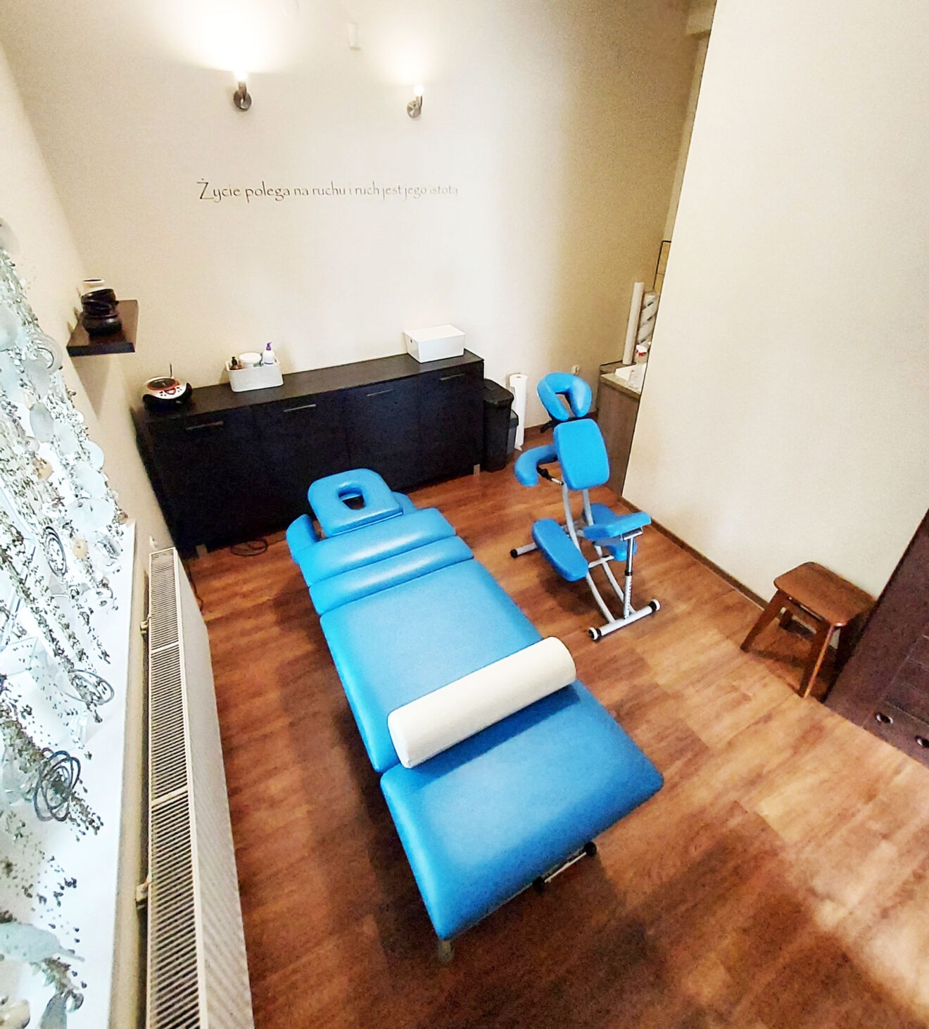 Jasne wnętrze gabinetu z niebieskim stołem do masażu z białym wałkiem, krzesłem do masażu na metalowej ramie, drewnianym stołkiem i szafką, na ścianie napis 'Życie polega na ruchu i ruch jest jego...