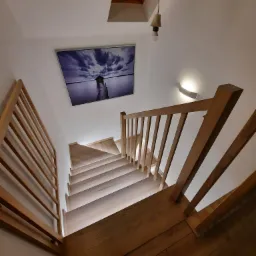 Drewniane schody z jasnego drewna z poręczą z tego samego materiału, oświetlone od spodu, widok z góry na dół, na ścianie obraz z motywem jeziora i chmur.