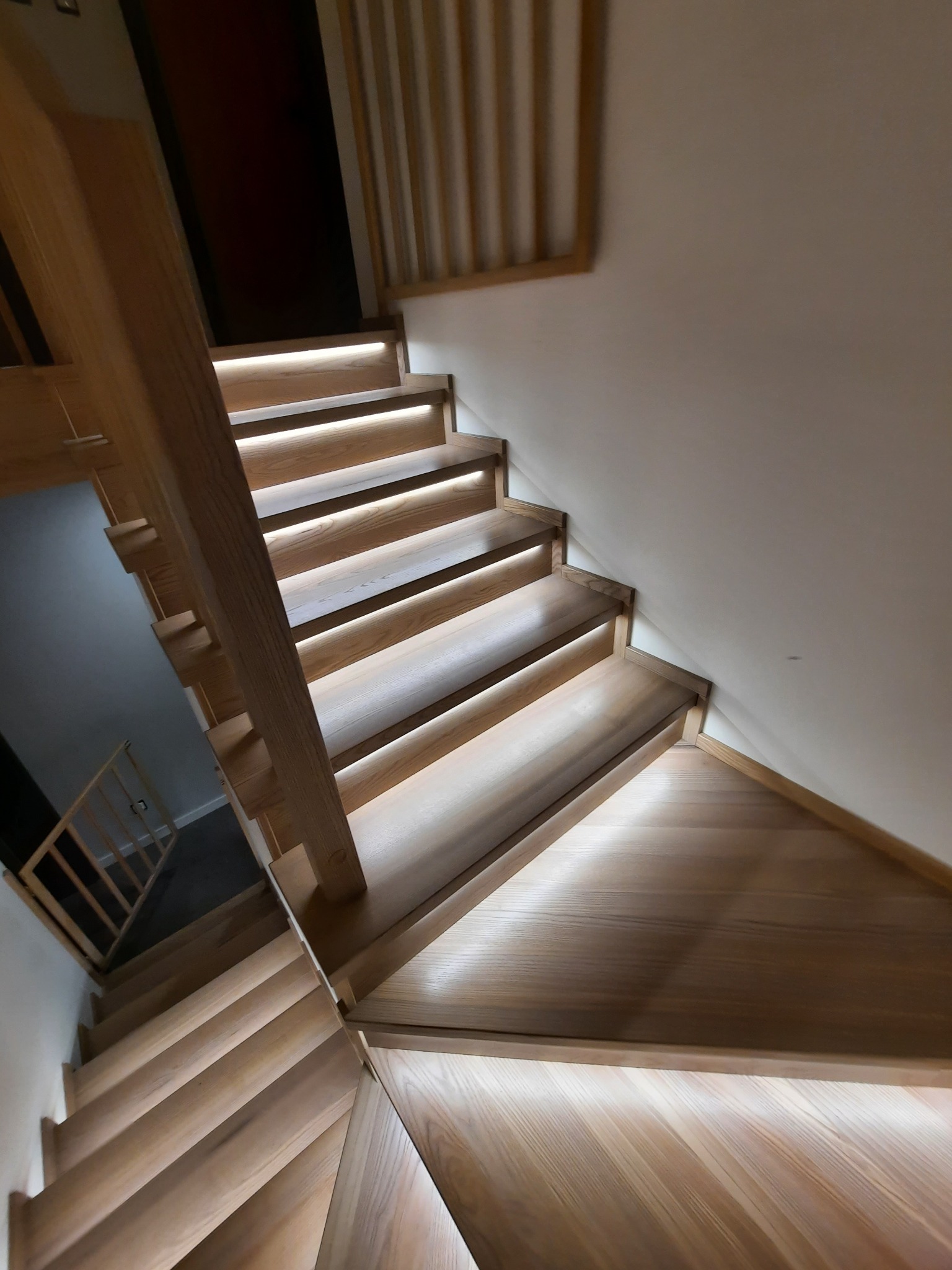 Jasne, drewniane schody z podświetleniem LED pod każdym stopniem, widziane z góry pod kątem, z widoczną balustradą na dole.