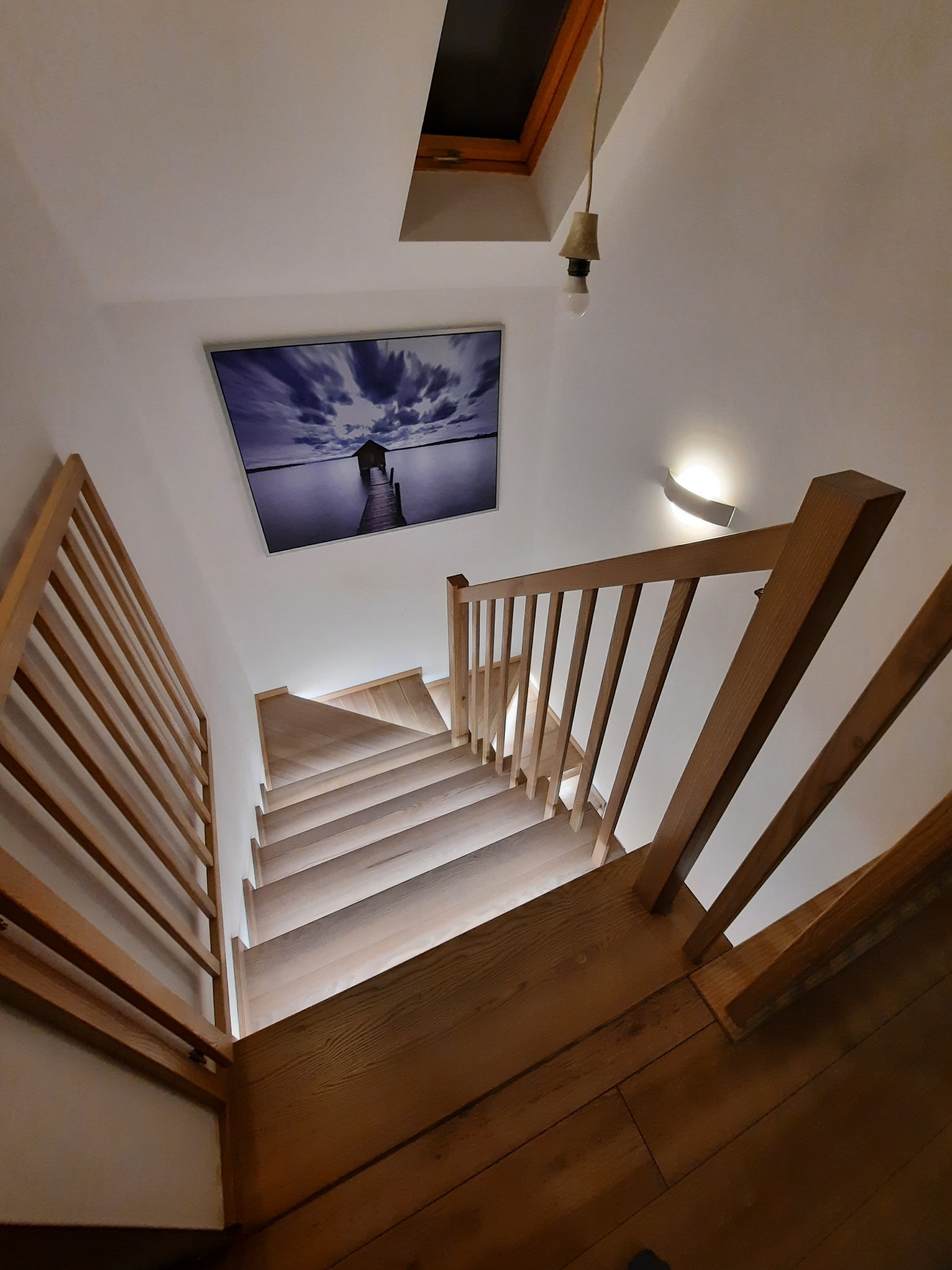 Drewniane schody z jasnego drewna z poręczą z tego samego materiału, oświetlone od spodu, widok z góry na dół, na ścianie obraz z motywem jeziora i chmur.