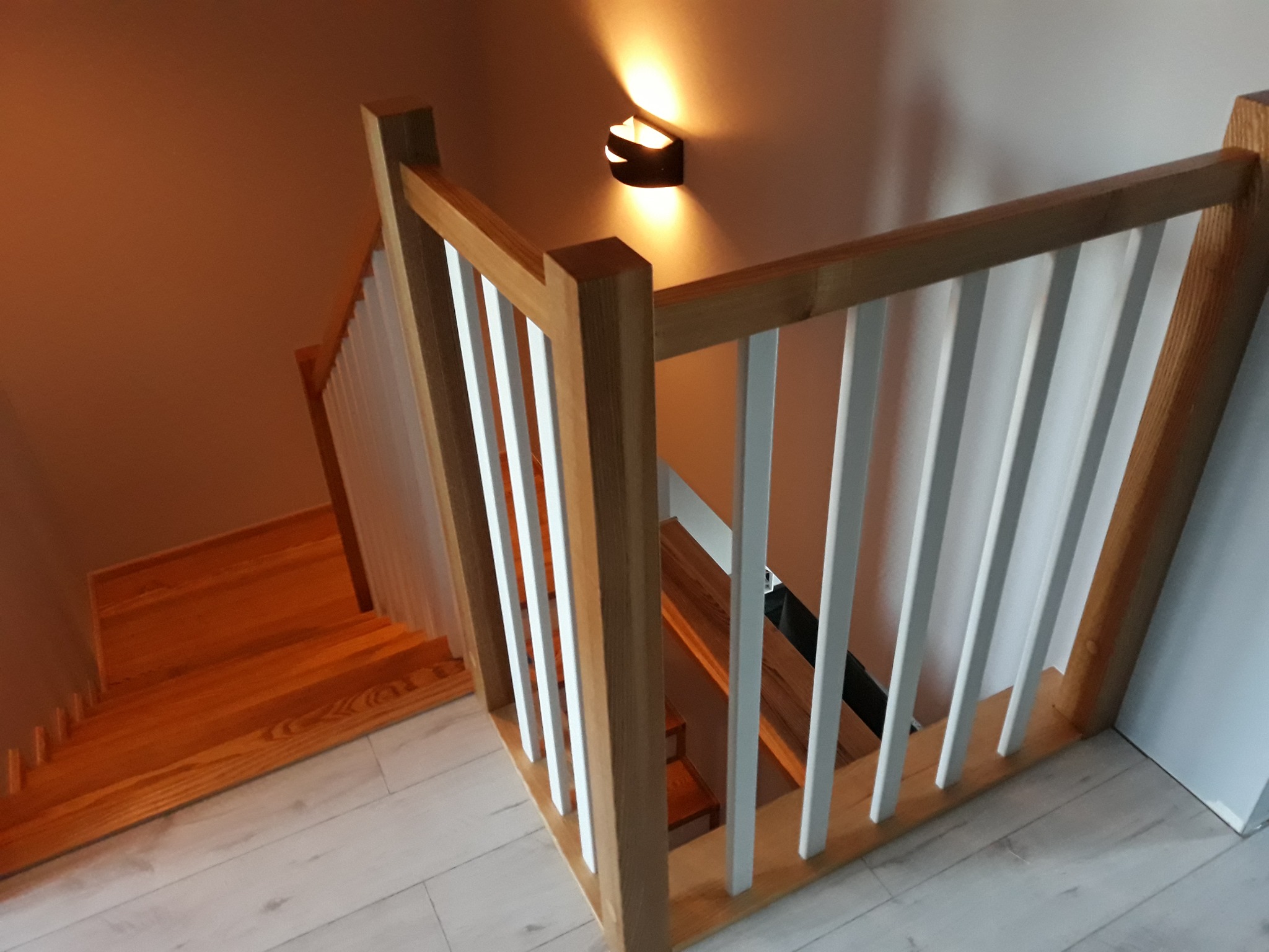 Widok z góry na drewniane schody z jasnymi poręczami w domu w Gwizdanowie, oświetlone nowoczesnym kinkietem.