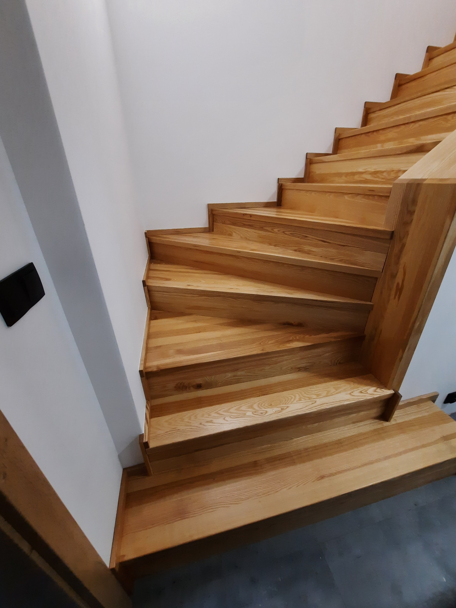 Nowoczesne, jasne stopnie z drewna z widocznym usłojeniem, prowadzące w górę wzdłuż białej ściany. Minimalistyczny design i brak balustrady.