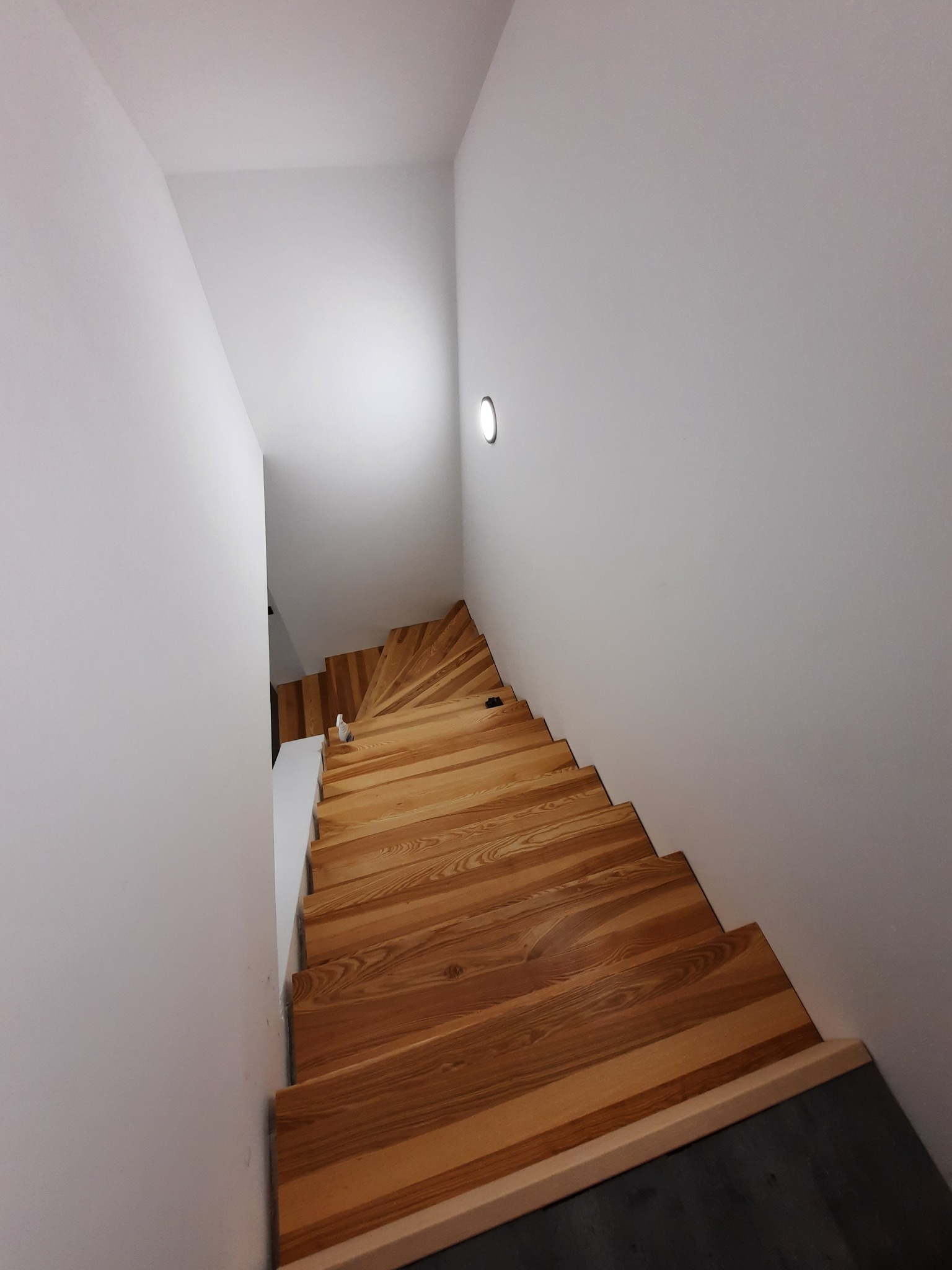Widok z góry na kręte schody z jasnego drewna, kontrastujące z białymi ścianami i ciemną podłogą na dole, oświetlone punktowym światłem.