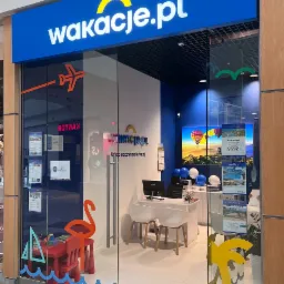 Wejście do biura podróży Wakacje.pl z logo nad drzwiami, z naklejkami na szybie przedstawiającymi flaminga, żaglówkę i palmę, oraz z widocznym wnętrzem z biurkiem, komputerem i dekoracjami.