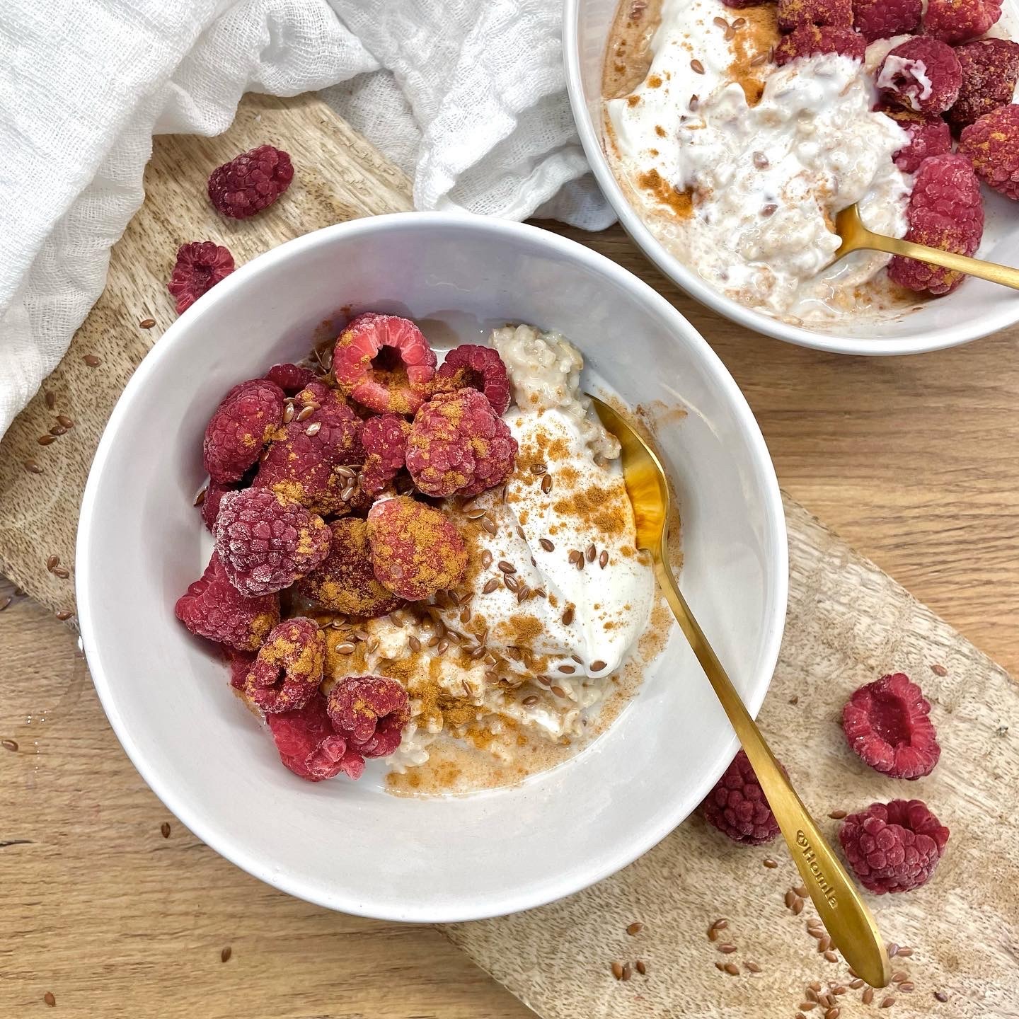 Dwie miseczki owsianki z jogurtem, malinami i siemieniem lnianym na drewnianej desce, złota łyżka w jednej z miseczek.