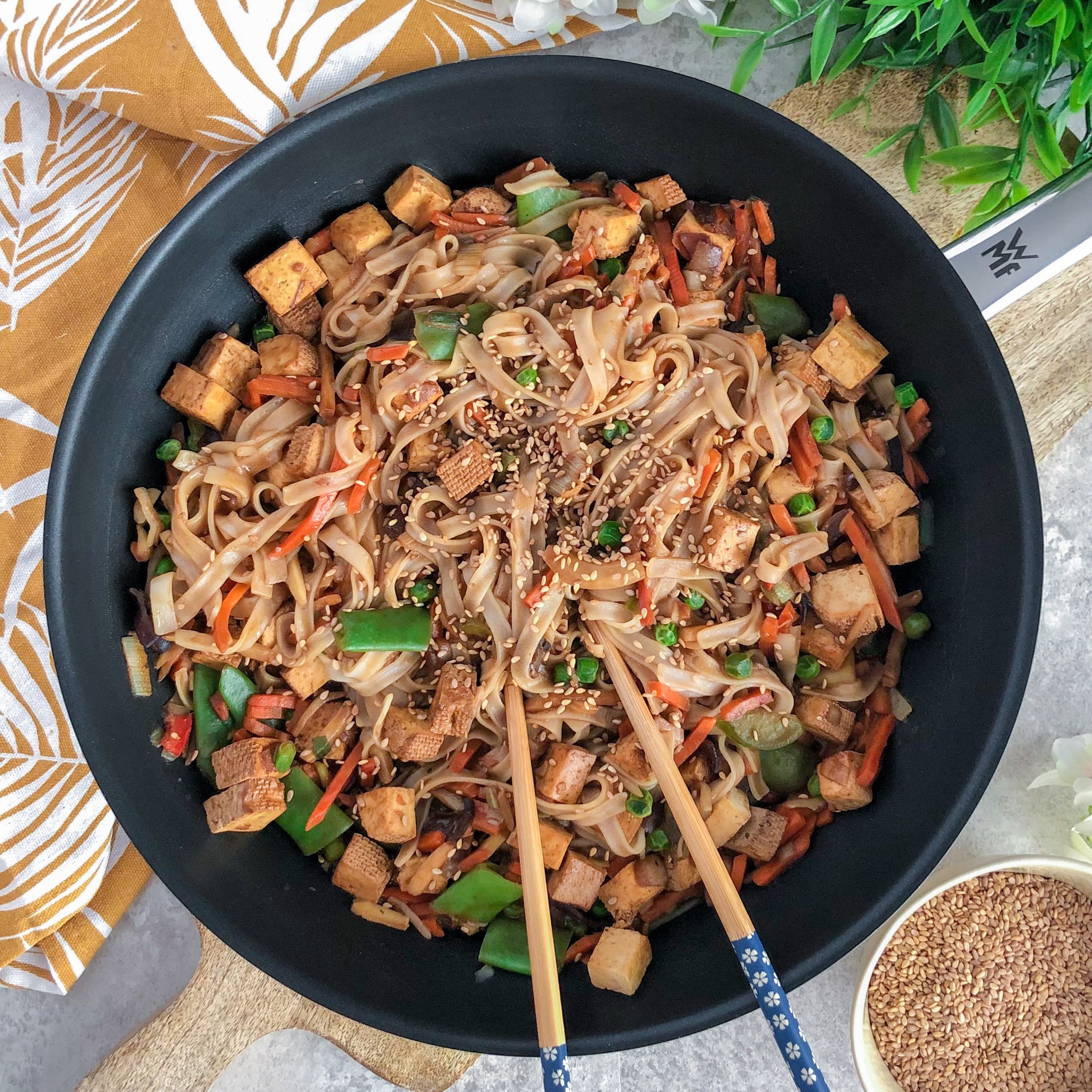 Wok z makaronem ryżowym, tofu, marchewką, groszkiem, papryką i sezamem, podawany z pałeczkami na drewnianej desce.