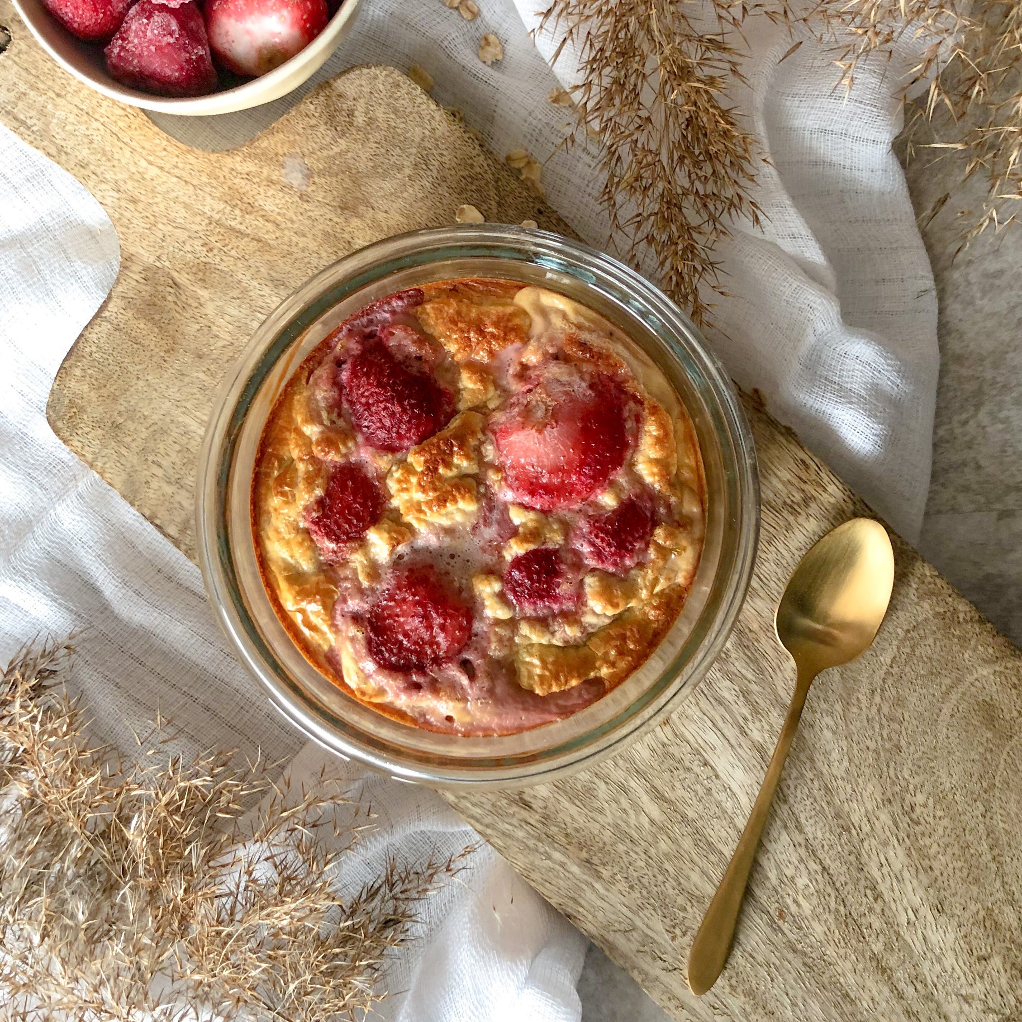 Zapiekanka owsiana z truskawkami w szklanym naczyniu, leżąca na drewnianej desce obok złotej łyżeczki, z miseczką mrożonych truskawek i dekoracją z suszonych traw w tle.