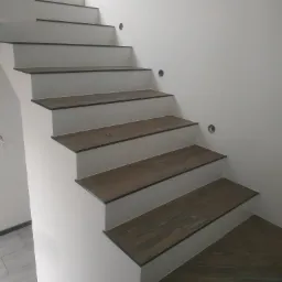 Wykończone schody z drewnopodobnymi płytkami na stopniach i białymi podstopnicami, widoczne otwory na punkty świetlne w ścianie.