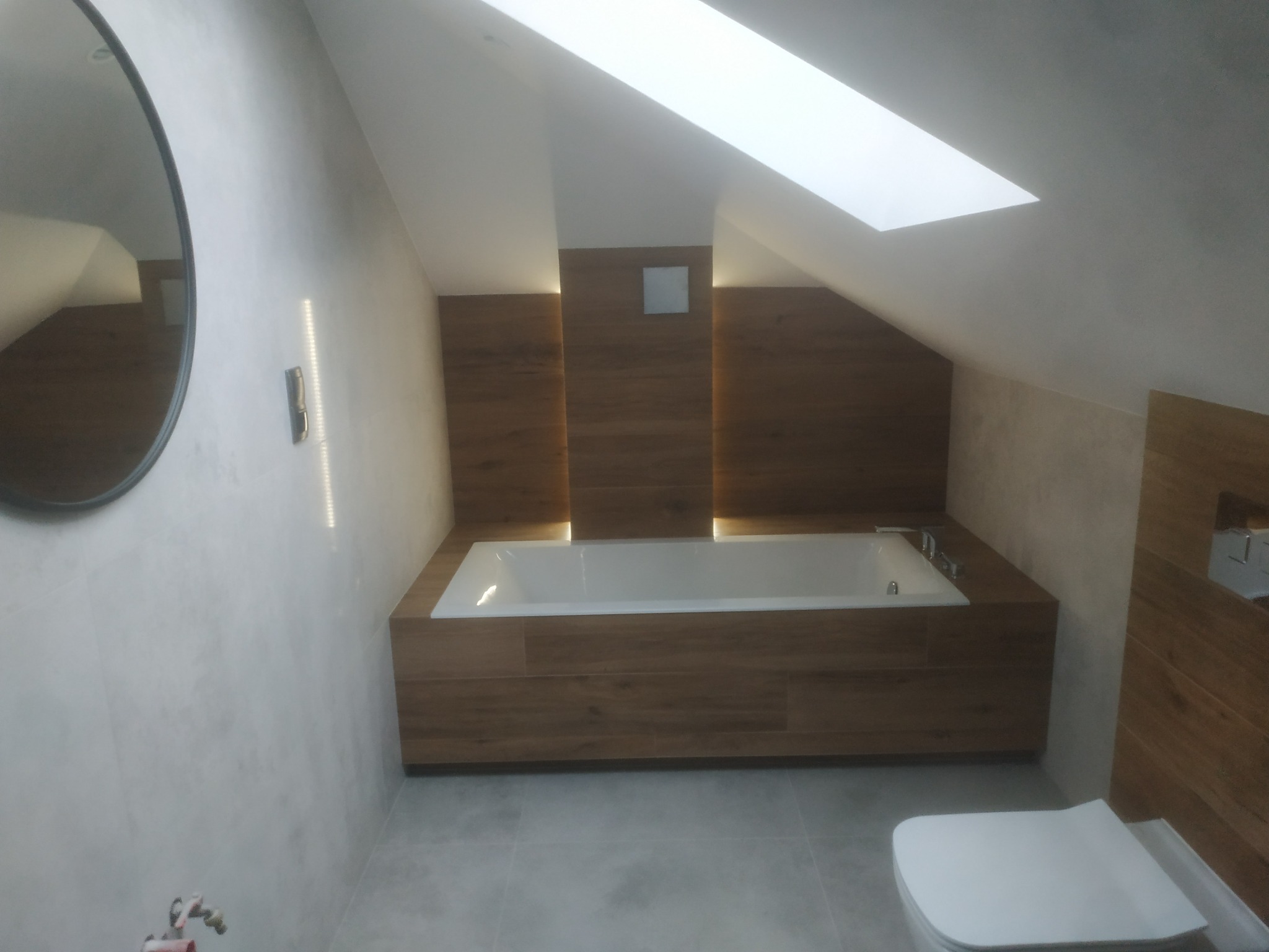 Nowoczesna łazienka na poddaszu z oknem dachowym, wykończona płytkami imitującymi drewno, z owalnym lustrem i podświetlaną wanną.