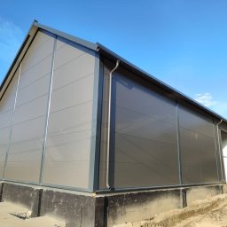 Automat Rent Sp.z.o.o. - Hala z płyt warstwowych w odcieniach szarości, widoczna z zewnątrz, z wyraźnymi panelami i elementami konstrukcyjnymi. W tle błękitne niebo i częściowo widoczne budynki.