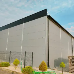 Nowoczesna hala z szarej płyty warstwowej, czarny dach, ogrodzenie z zielonymi krzewami. Widok z zewnątrz na tle błękitnego nieba.