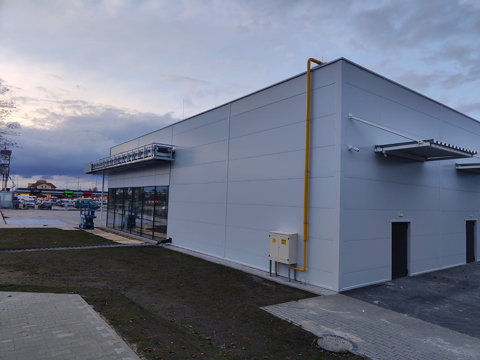 Nowoczesna hala z płyt warstwowych w kolorze szarym, z widoczną żółtą rurą gazową i elementami konstrukcyjnymi. Ujęcie z zewnątrz, z fragmentem parkingu w tle.