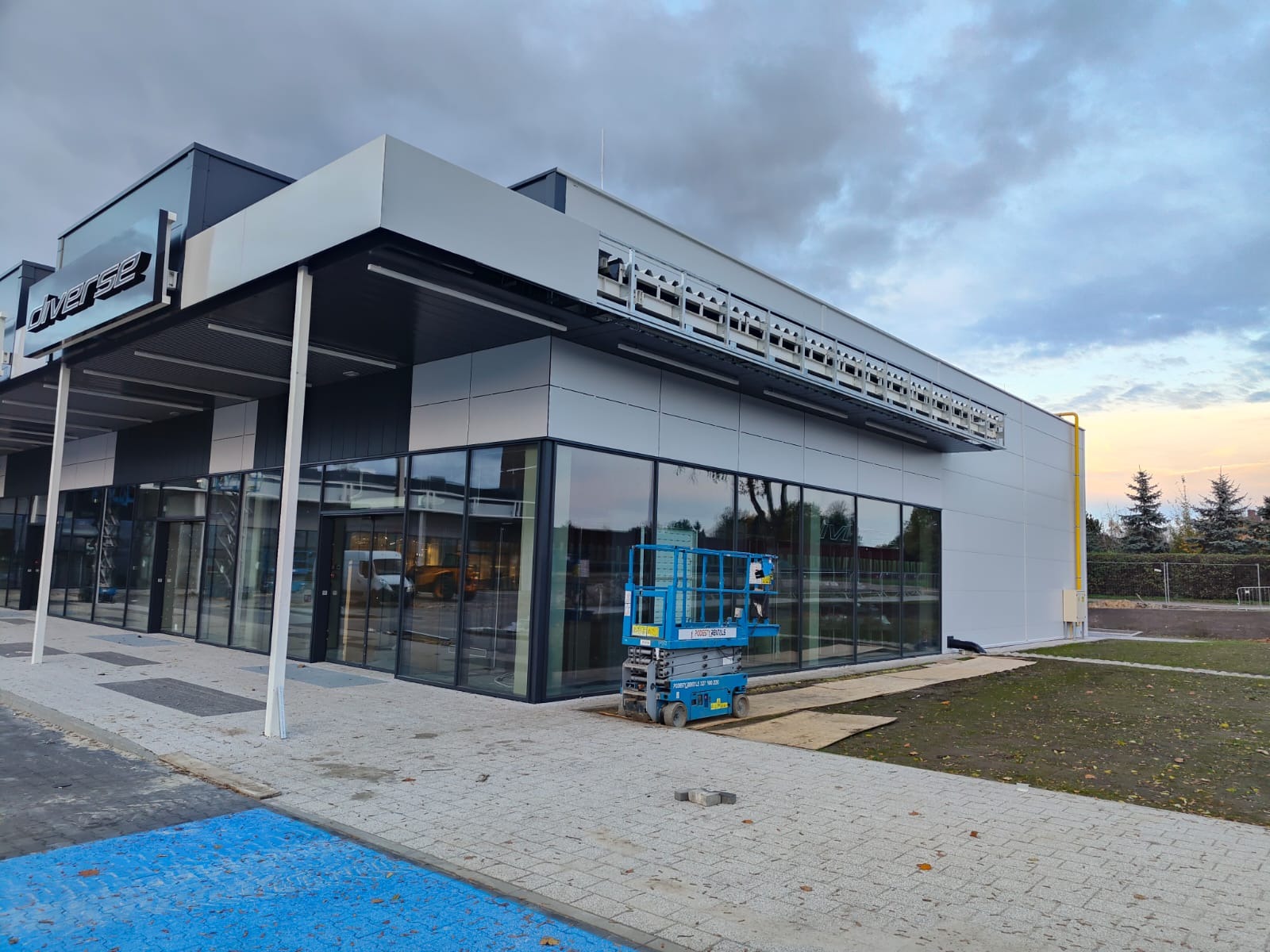 Nowoczesna hala handlowa z płyt warstwowych, widoczna podnośnik koszowy. Elewacja w odcieniach szarości, duże okna. Ujęcie z zewnątrz, w tle pochmurne niebo.