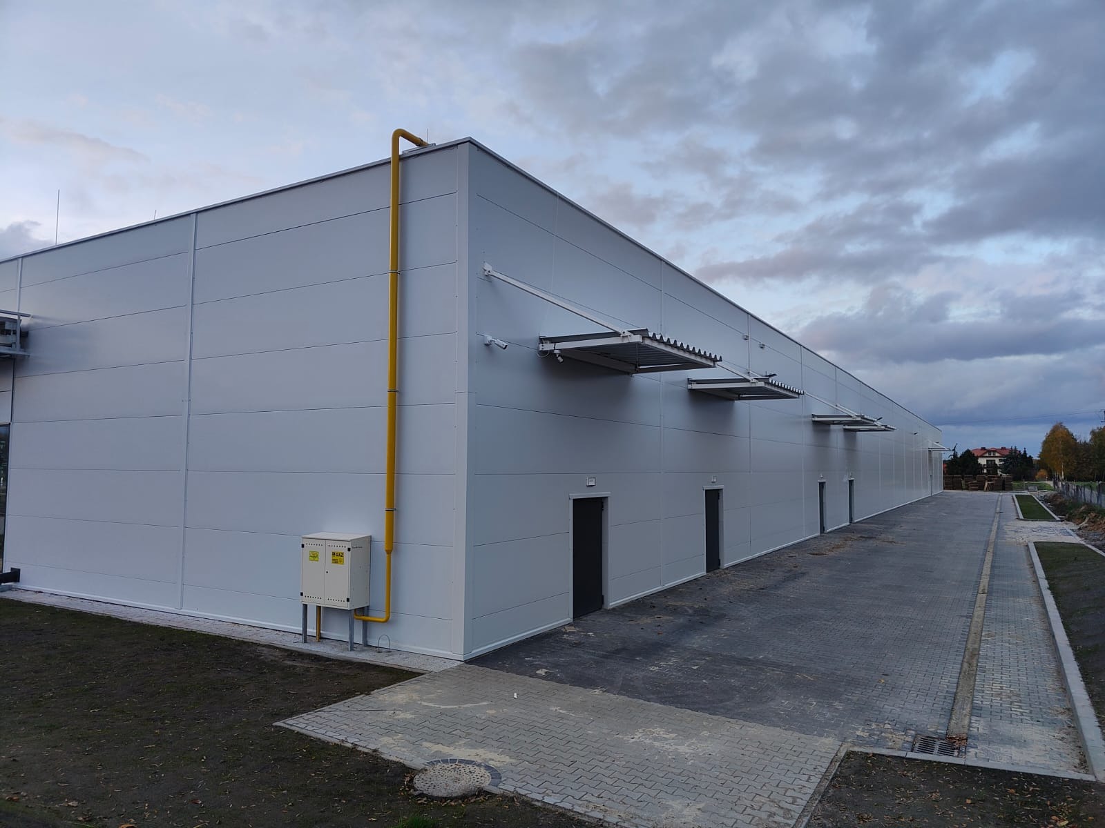 Nowoczesna hala z płyt warstwowych, widok z zewnątrz. Żółta rura gazowa na bocznej ścianie, szare drzwi wejściowe, metalowe daszki nad wejściami. Pochmurne niebo w tle.