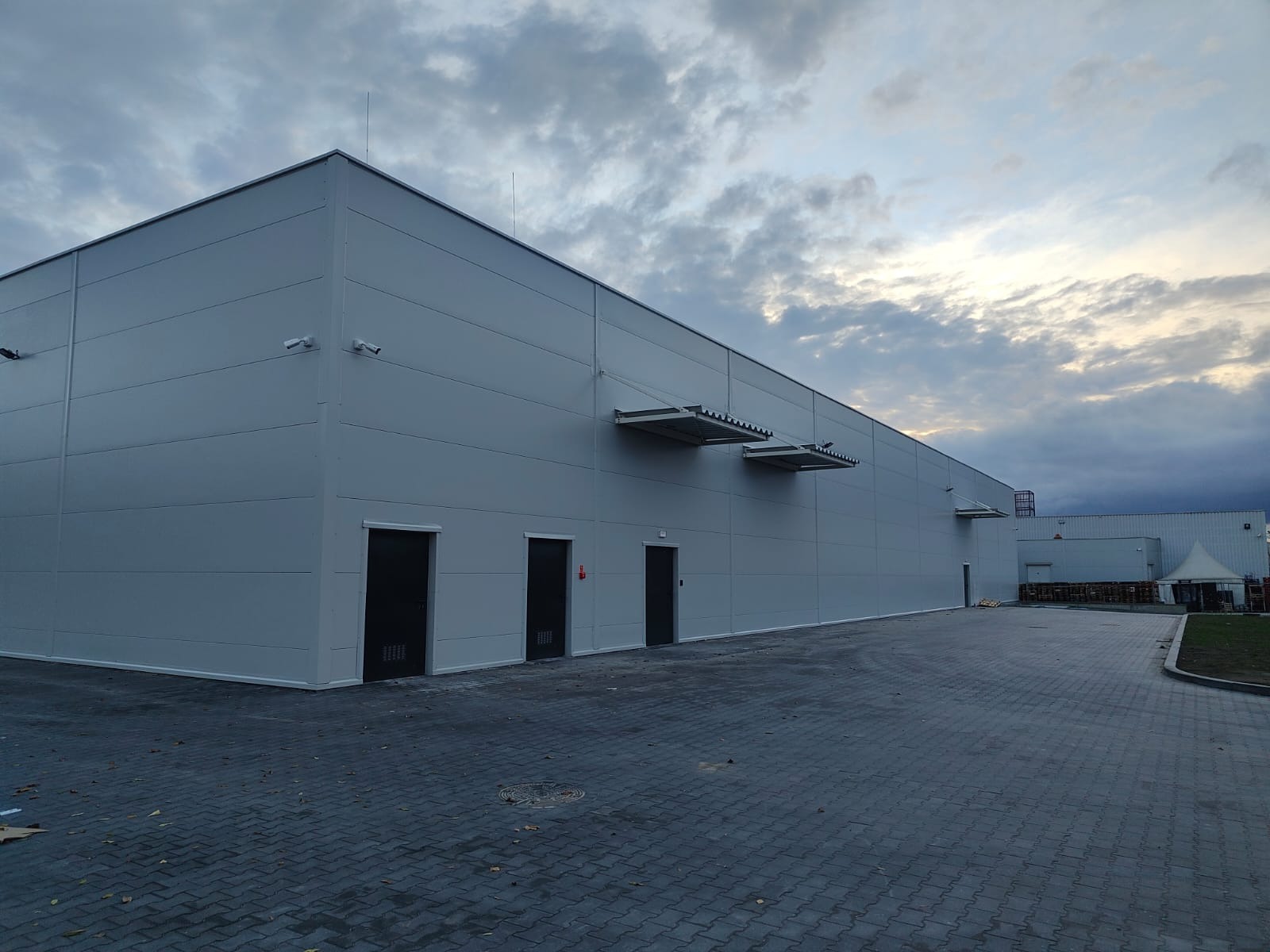 Nowoczesna hala przemysłowa z szarych płyt warstwowych, widok z zewnątrz. Widoczne drzwi, kamery monitoringu i zadaszenia nad wejściami. Pochmurne niebo w tle.