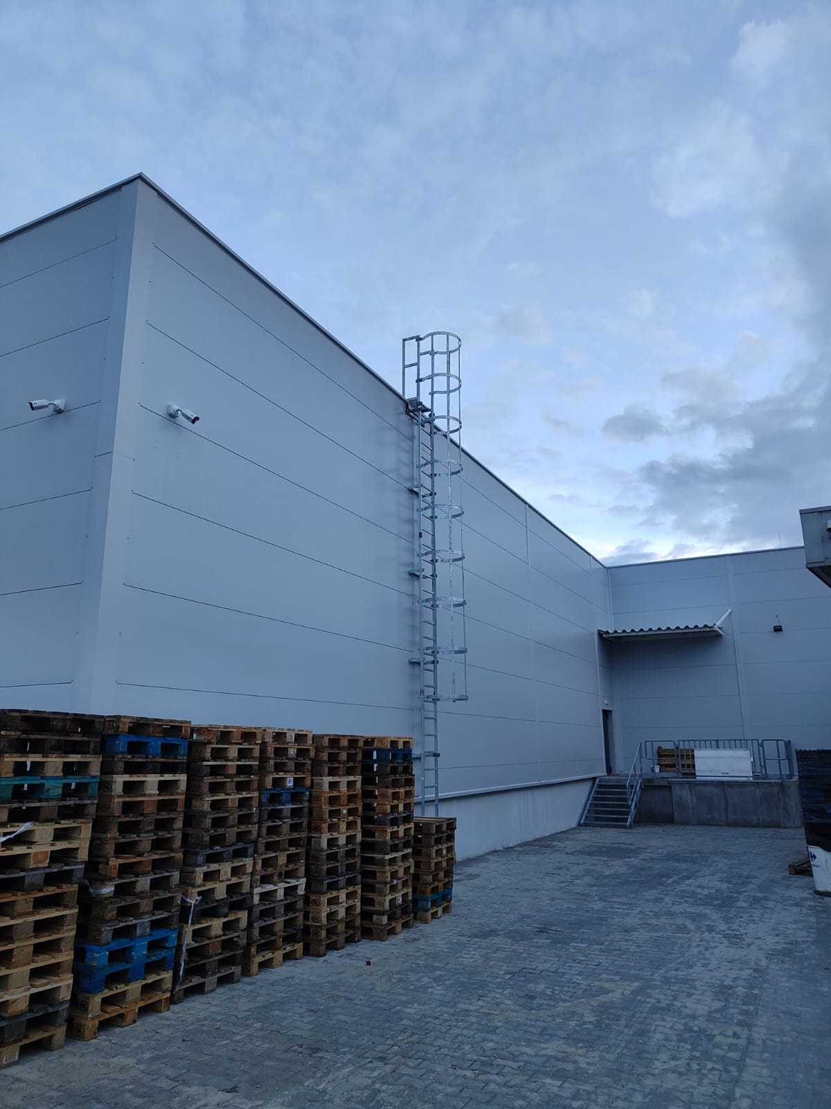 Hala z płyt warstwowych, z drabinką na elewacji i kamerami przemysłowymi. Stos palet w dolnej części kadru. Pochmurne niebo w tle.