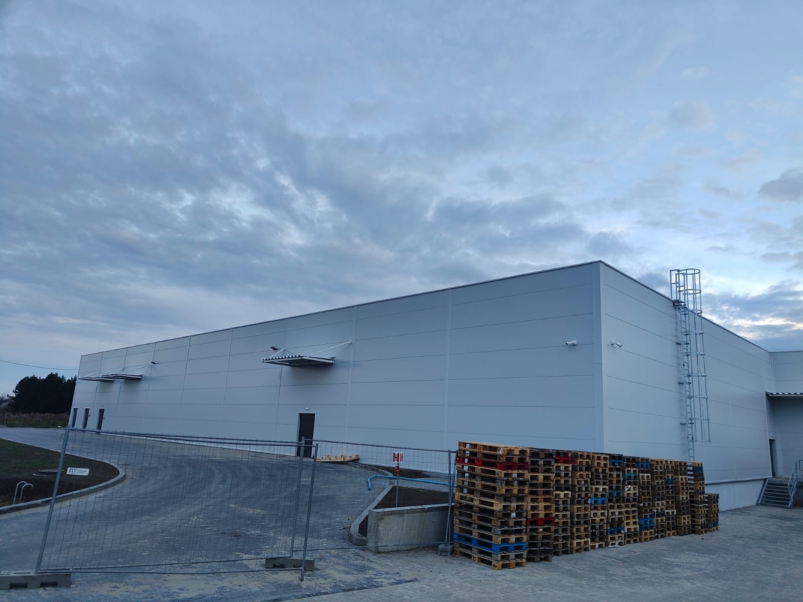 Nowoczesna hala magazynowa z szarej płyty warstwowej, widok z zewnątrz. Na pierwszym planie palety, w tle pochmurne niebo. Widoczna drabina na ścianie.