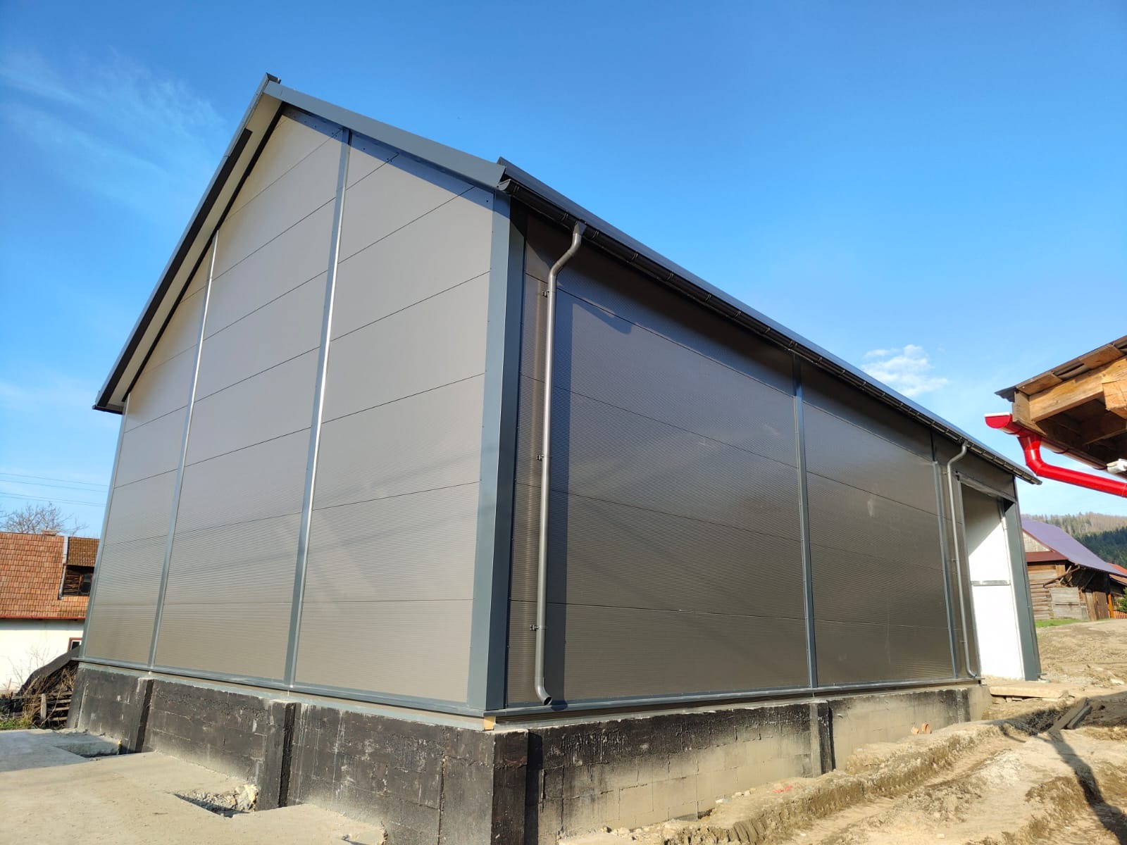 Hala z płyt warstwowych w odcieniach szarości, widoczna z zewnątrz, z wyraźnymi panelami i elementami konstrukcyjnymi. W tle błękitne niebo i częściowo widoczne budynki.