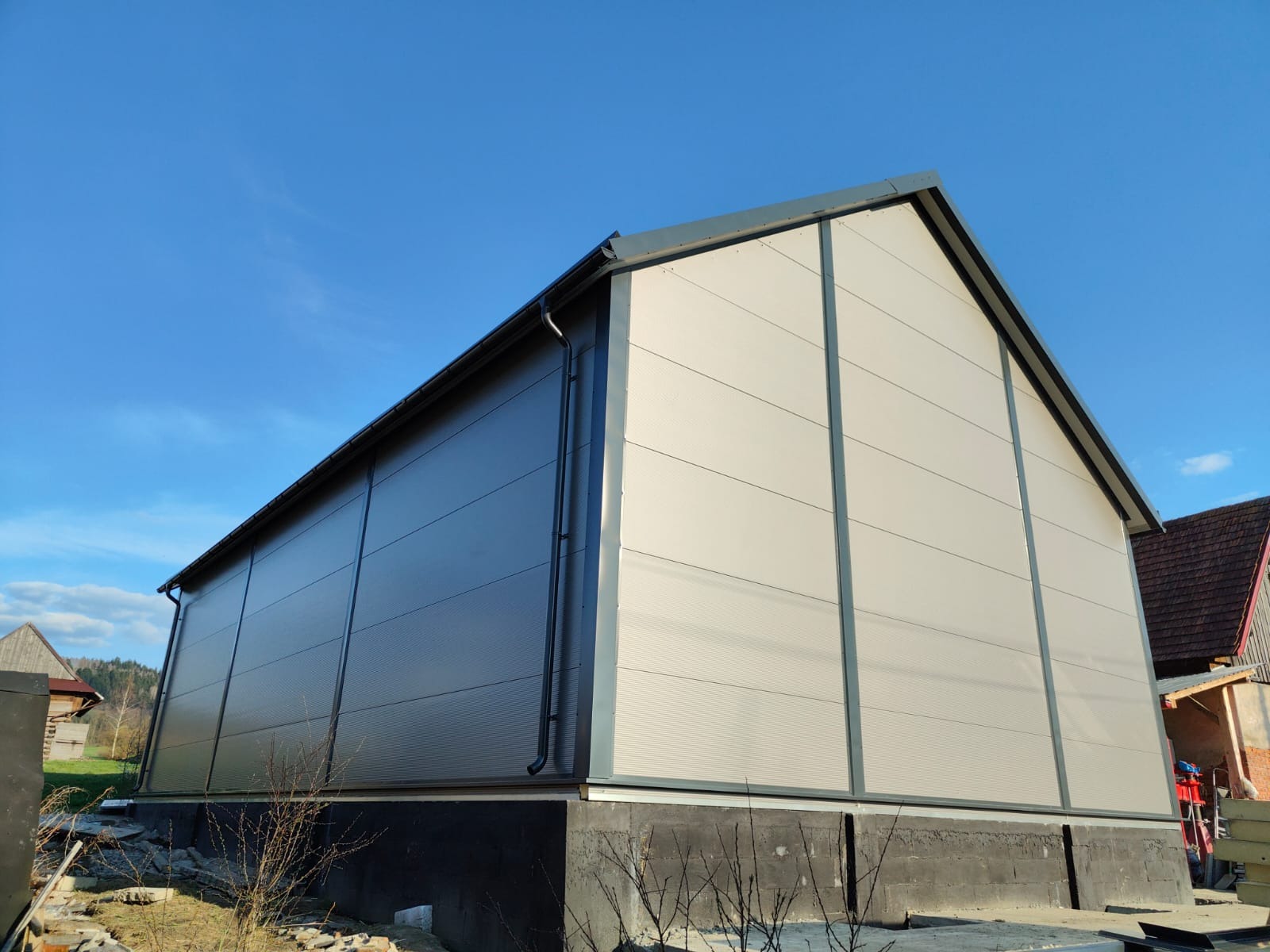 Nowoczesna hala z płyt warstwowych w odcieniach szarości, z widocznym systemem rynnowym, na tle błękitnego nieba i zabudowań wiejskich. Ujęcie z dołu podkreśla jej gabaryty.