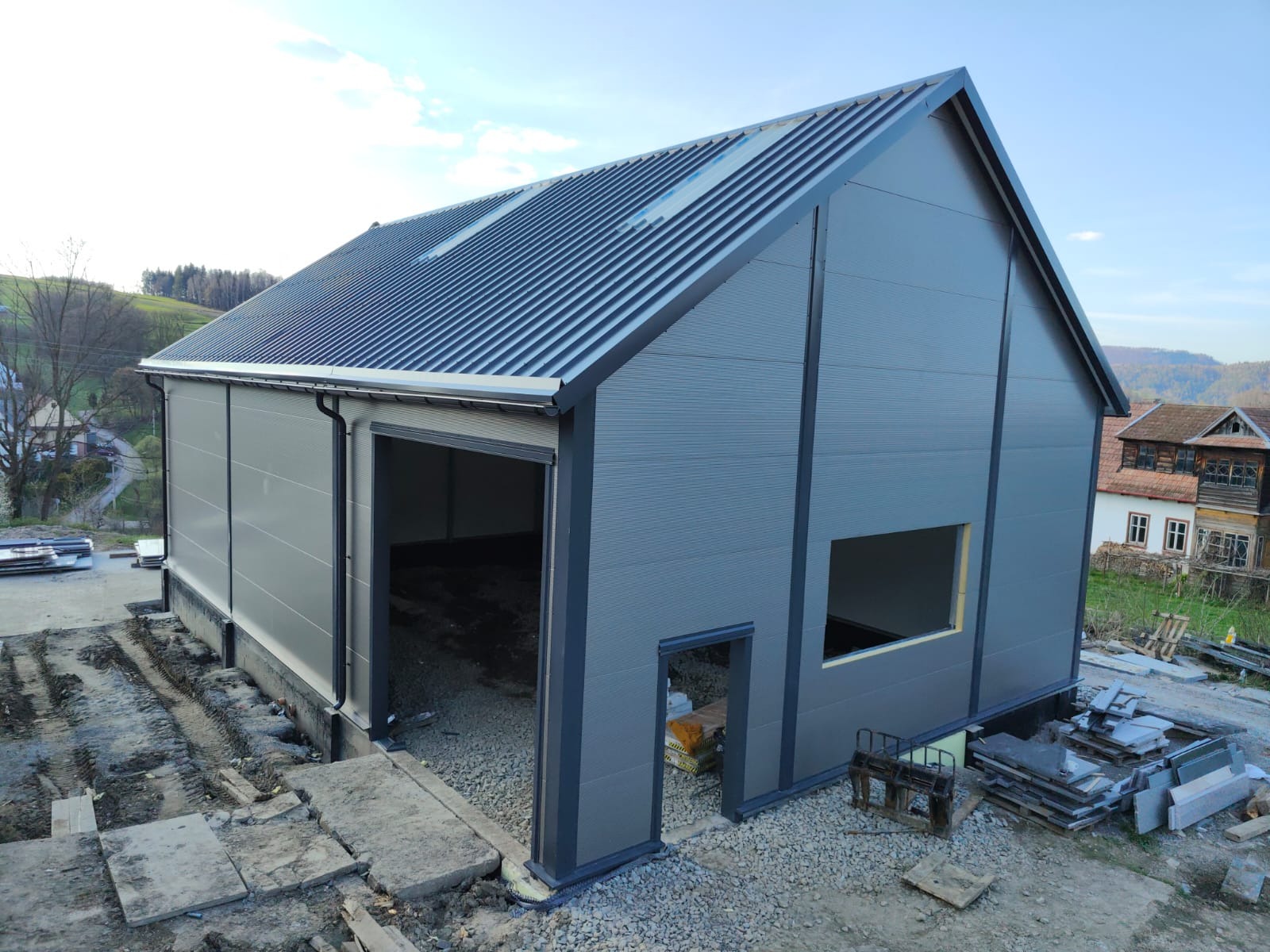 Szara hala z płyty warstwowej w trakcie budowy, widoczne otwory na okna i drzwi, dach z blachy trapezowej, teren budowy w nieładzie, w tle budynki mieszkalne.